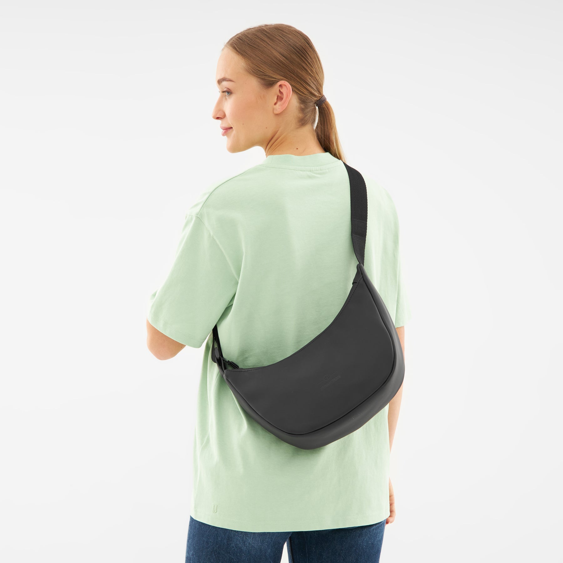 Frau mit einem schwarzen Half Moon Bag 'Rory' von Johnny Urban über der Schulter, trägt hellgrünes T-Shirt und blaue Jeans