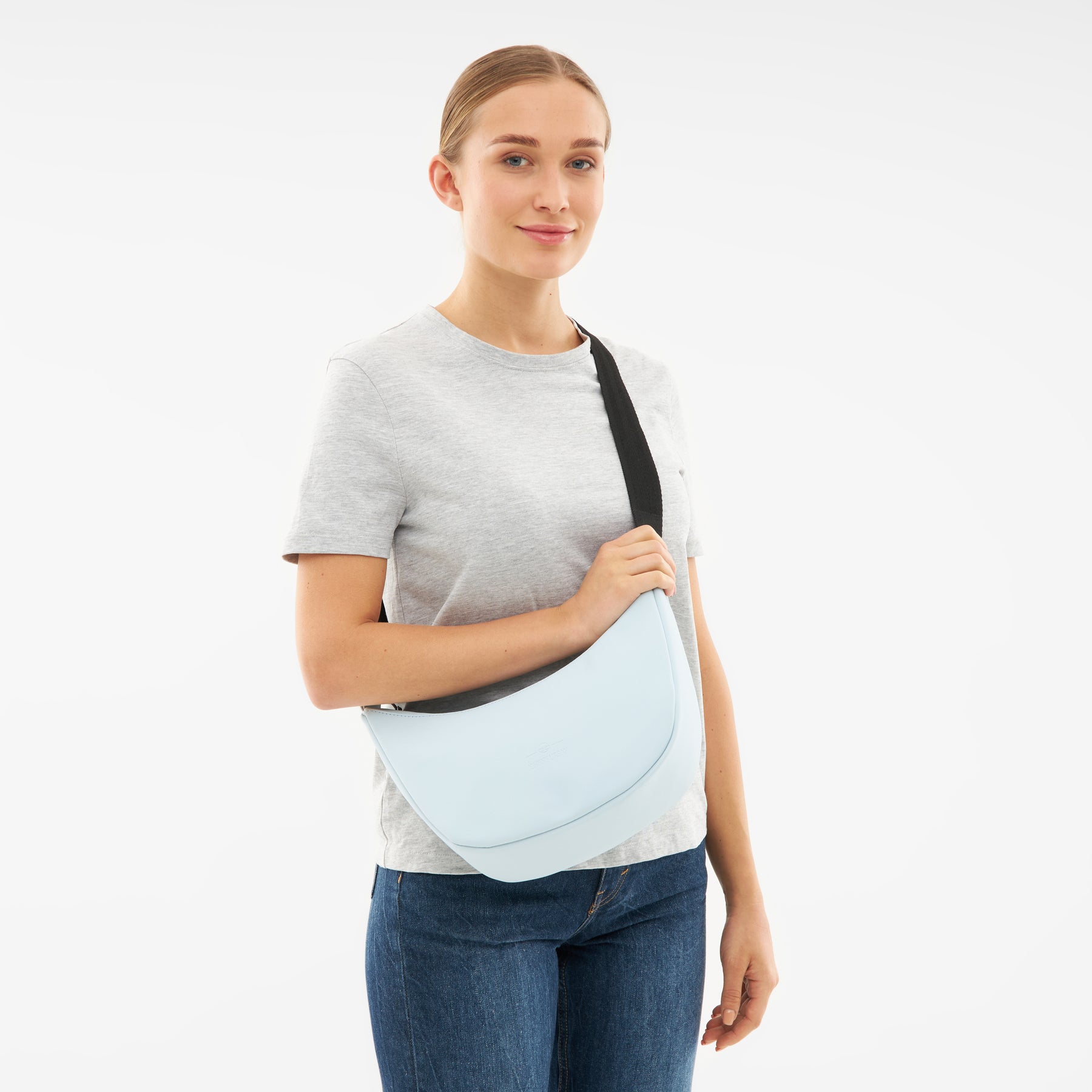 Frau trägt hellblaue Half Moon Bag 'Rory' mit schwarzem Tragegurt, graues T-Shirt und blaue Jeans