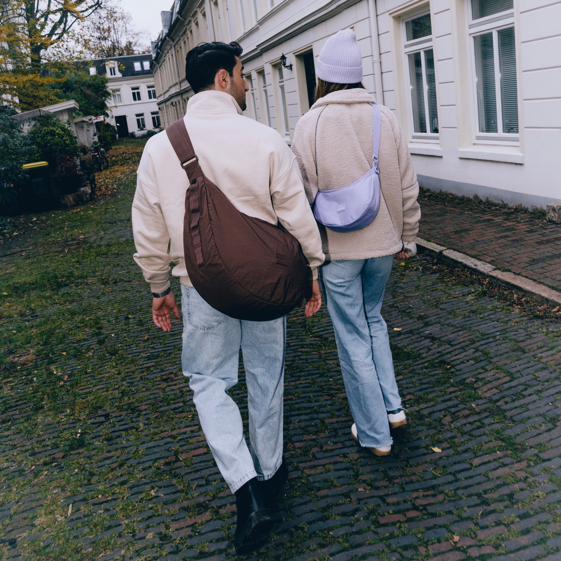 Zwei Personen gehen auf Kopfsteinpflaster, eine trägt die große braune Half Moon Bag 'Kaia XL', die andere eine kleine lila Half Moon Bag