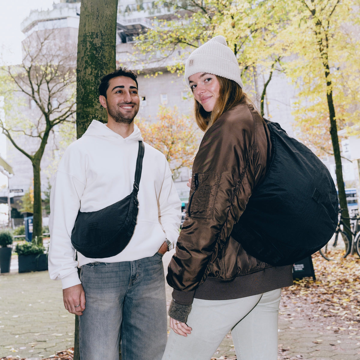 Mann mit hellbeigem Hoodie trägt schwarze Half Moon Bag Kaia XL crossbody, Frau mit brauner Jacke und weißer Mütze trägt sie als Rucksack