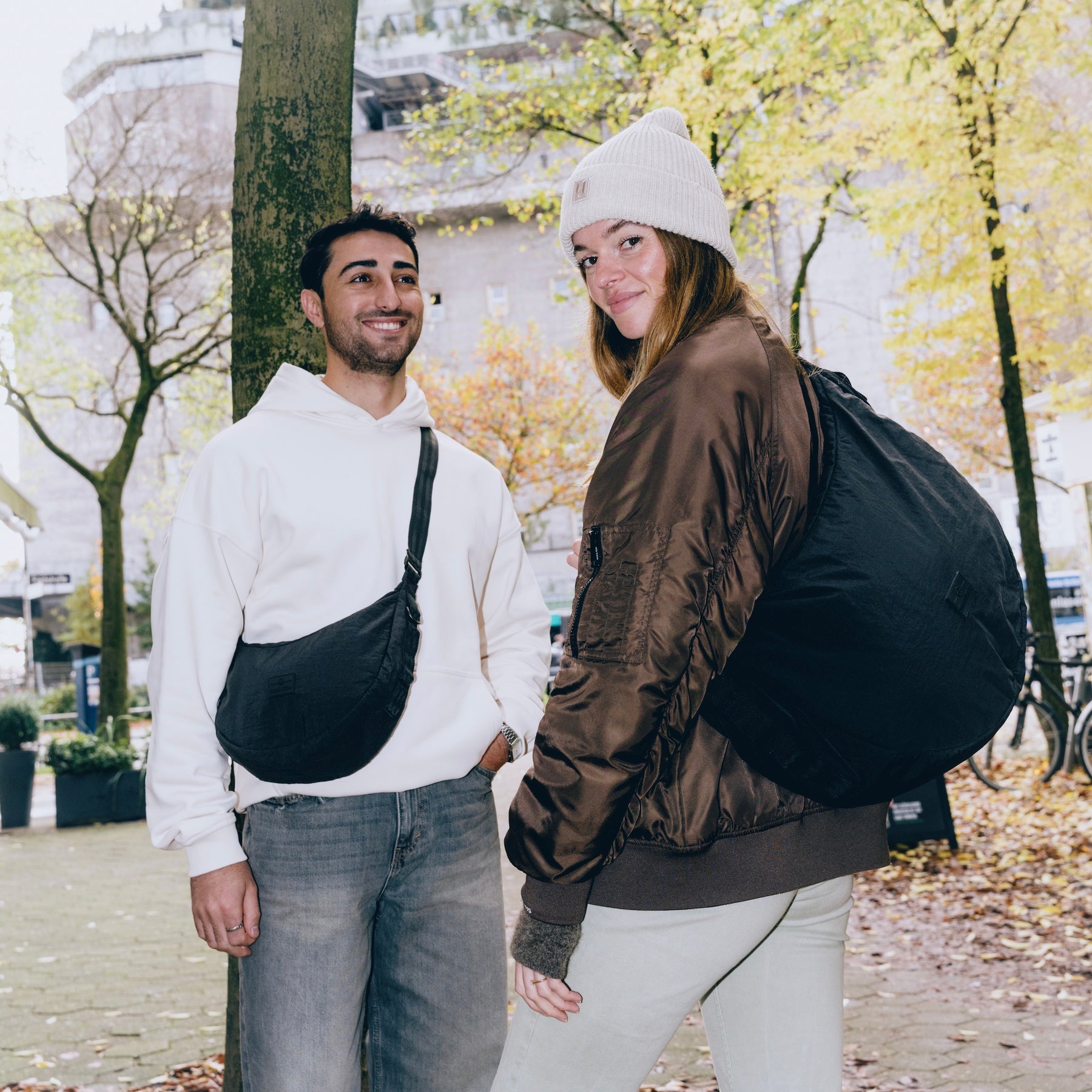 Mann mit hellbeigem Hoodie trägt schwarze Half Moon Bag Kaia XL crossbody, Frau mit brauner Jacke und weißer Mütze trägt sie als Rucksack