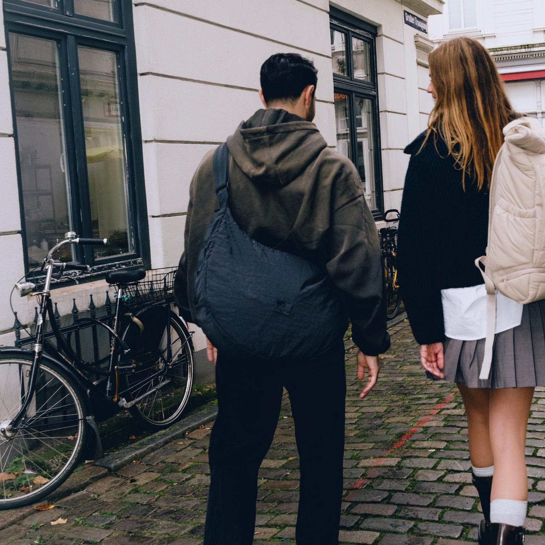 Mann trägt schwarze Half Moon Bag 'Kaia XL' von Johnny Urban über der Schulter, geht mit Frau auf Kopfsteinpflasterstraße