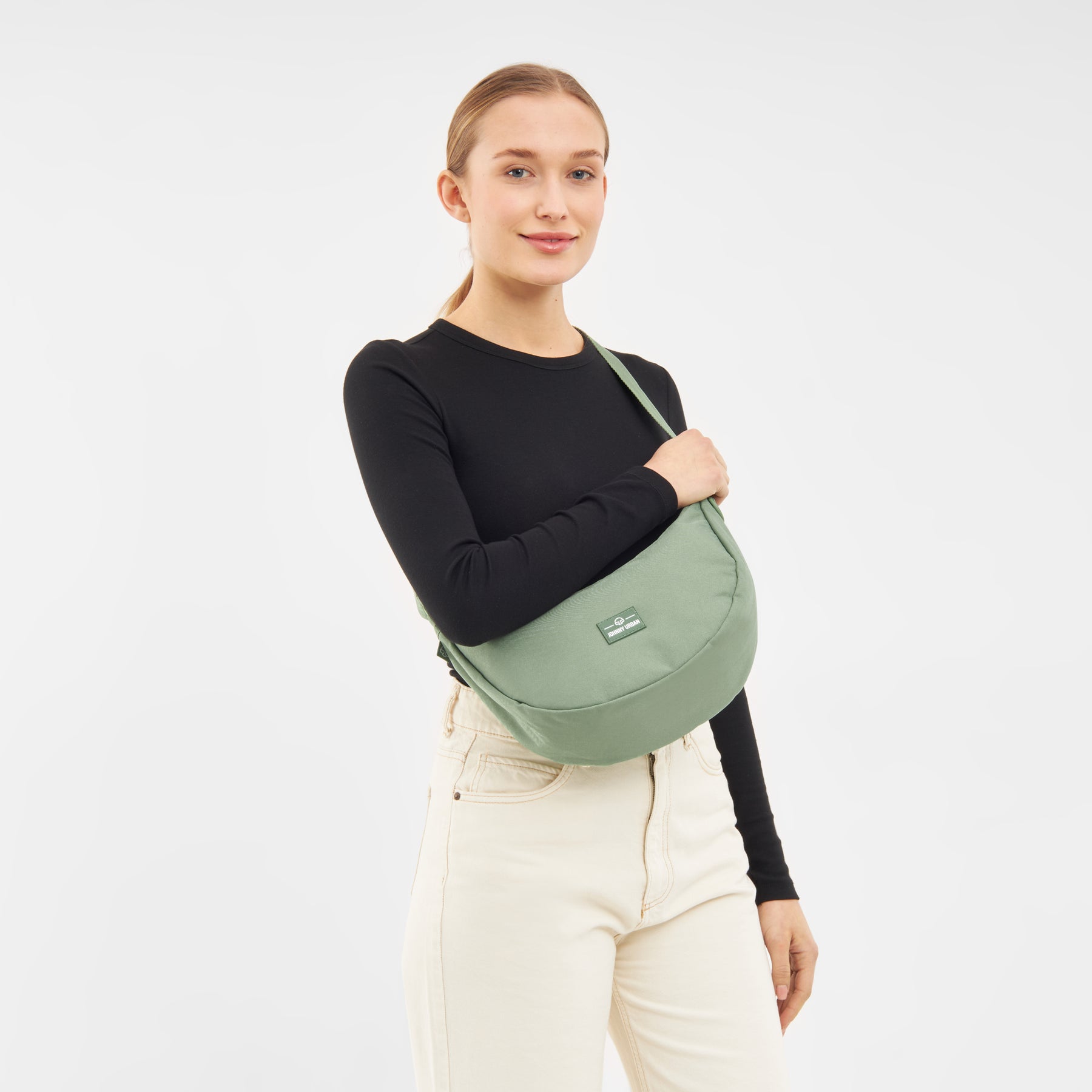 Frau mit halbmondenförmiger Tasche 'Ally Medium' in Salbeigrün, schwarzer langärmeliger Oberteil und helle Hose
