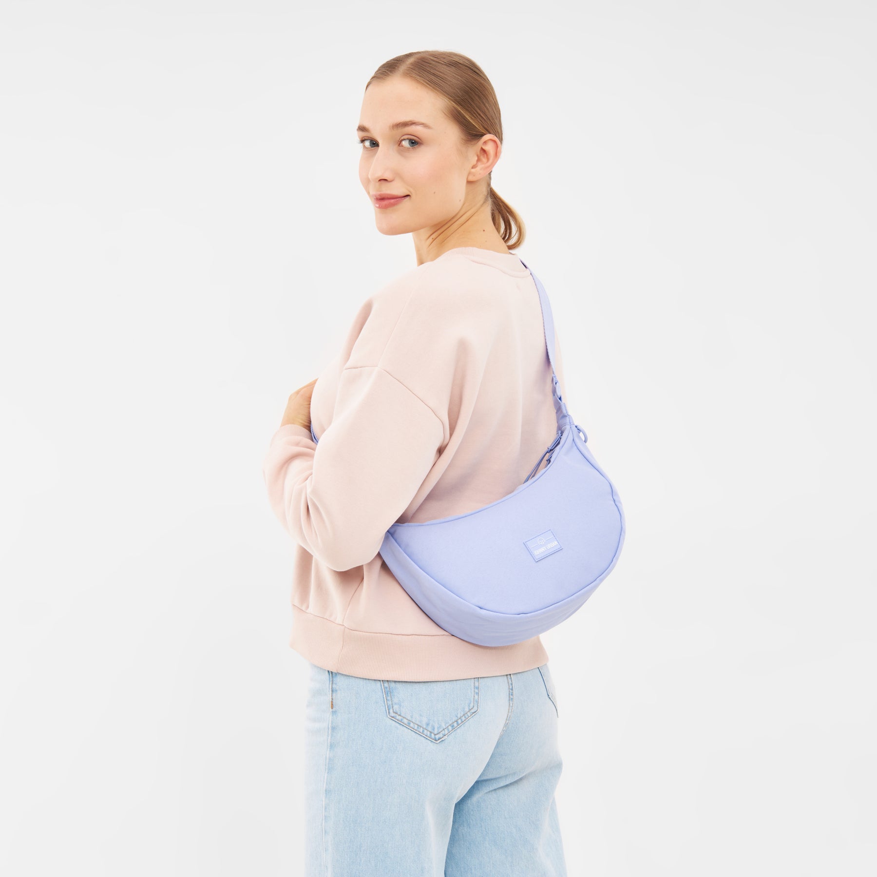 Frau trägt halbrunde Umhängetasche 'Ally Medium' in Hellblau über einer hellen Sweatshirtjacke und Jeans