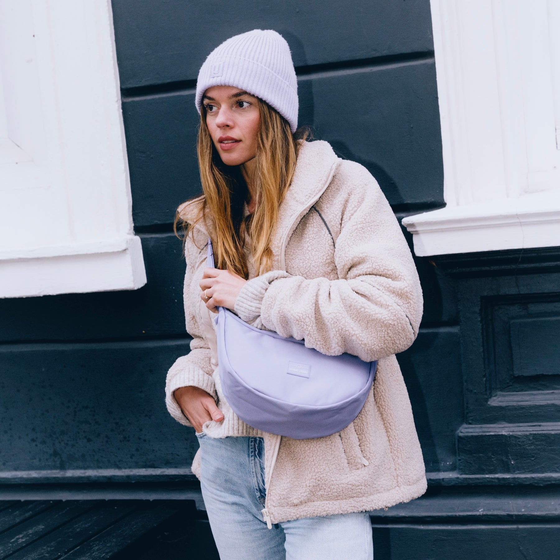 Frau mit hellviolettem Half Moon Bag 'Ally Medium', beiger Teddyjacke und Mütze vor schwarz-weißer Wand