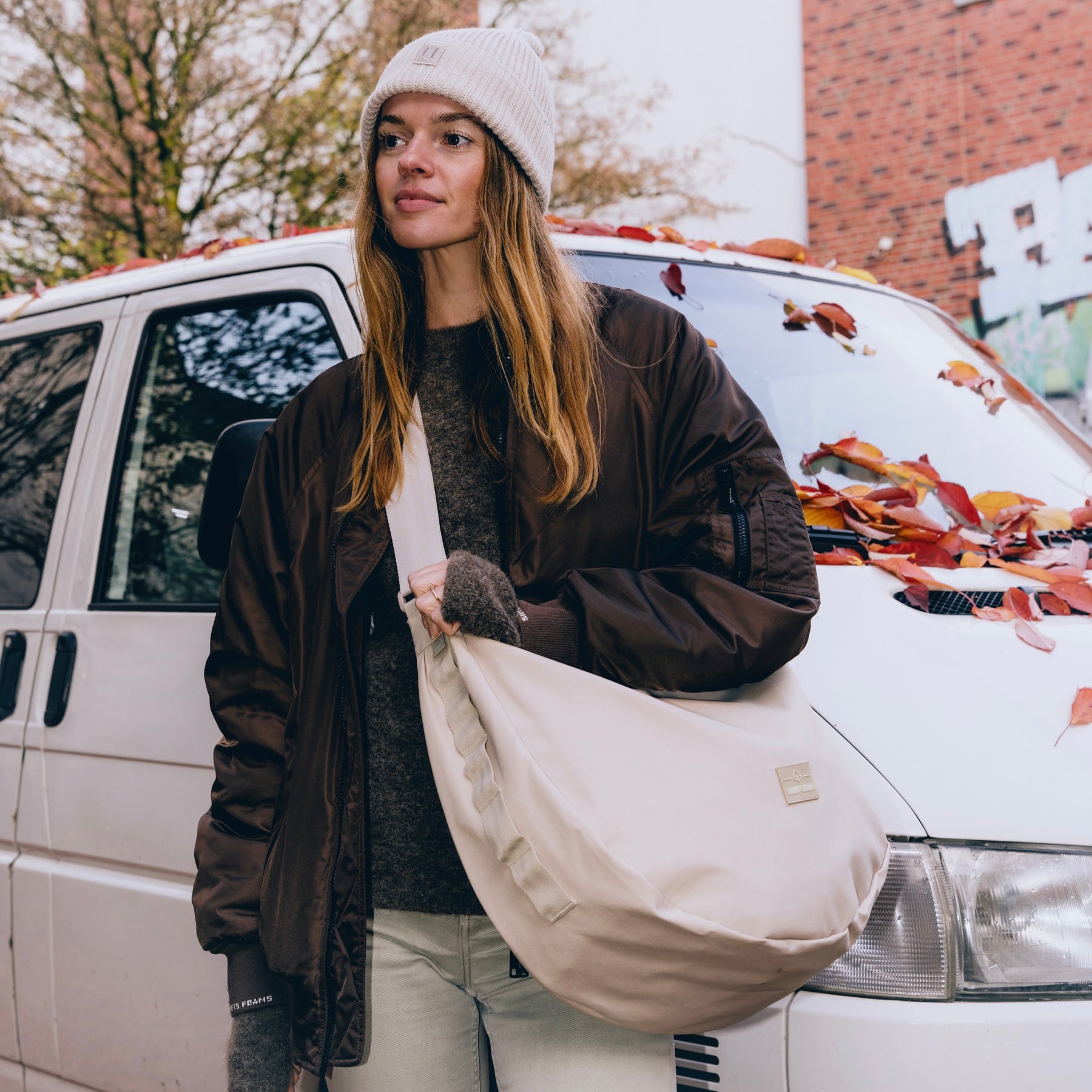 Frau mit halbrunder, hellbeiger Johnny Urban Umhängetasche 'Ally XL' vor weißem Van mit Herbstlaub