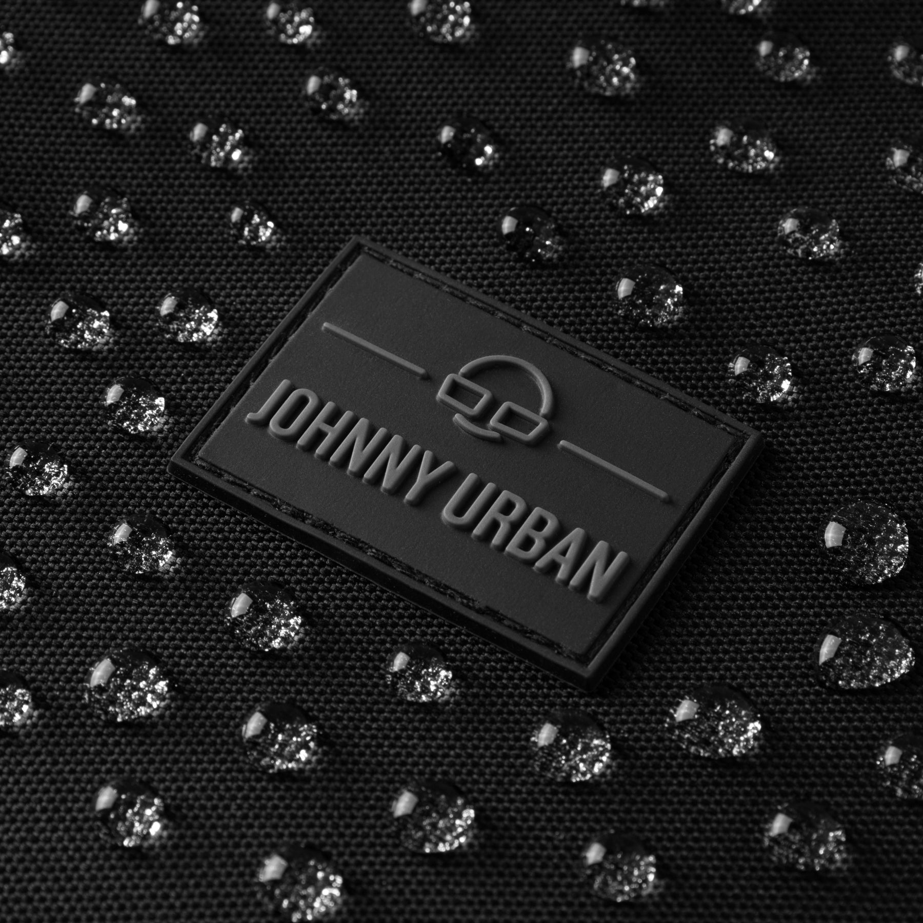 Nahe Ansicht eines schwarzen Johnny Urban Labels auf wasserabweisendem Material mit Wassertropfen auf der Halbmondtasche Ally XL