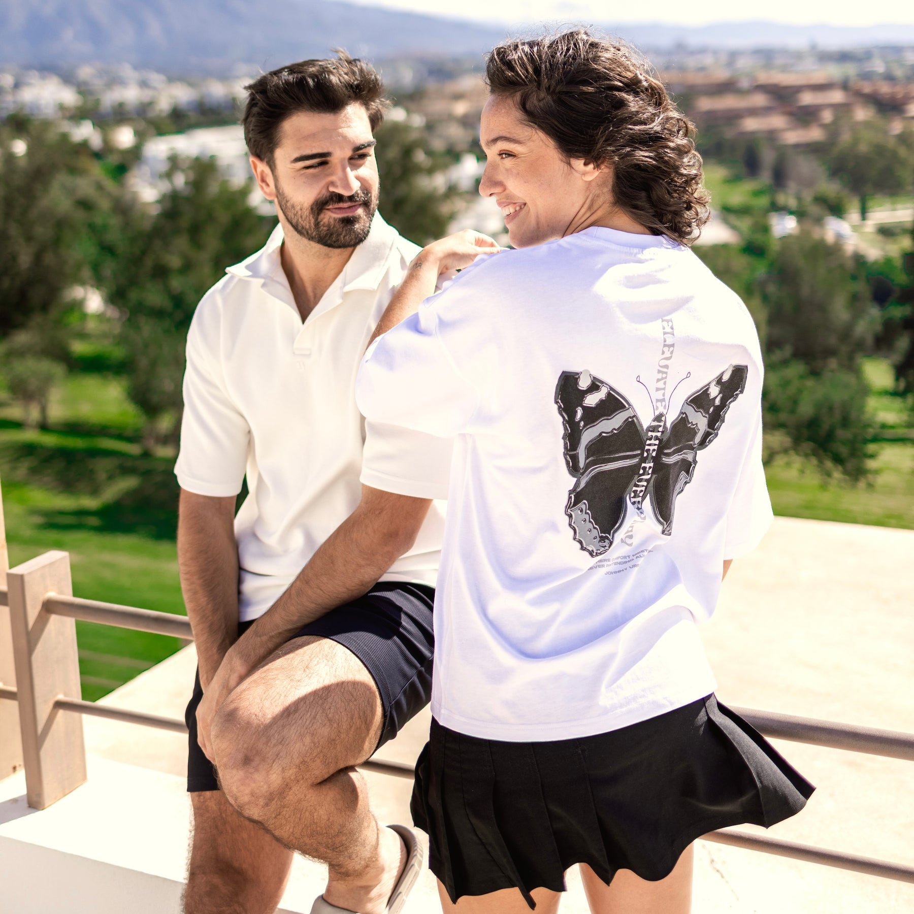 Frau trägt weißes Oversize-T-Shirt 'Sammy Chrome Butterfly' mit schwarzem Schmetterlingsmotiv auf dem Rücken, Mann daneben in weißem Poloshirt und schwarzen Shorts