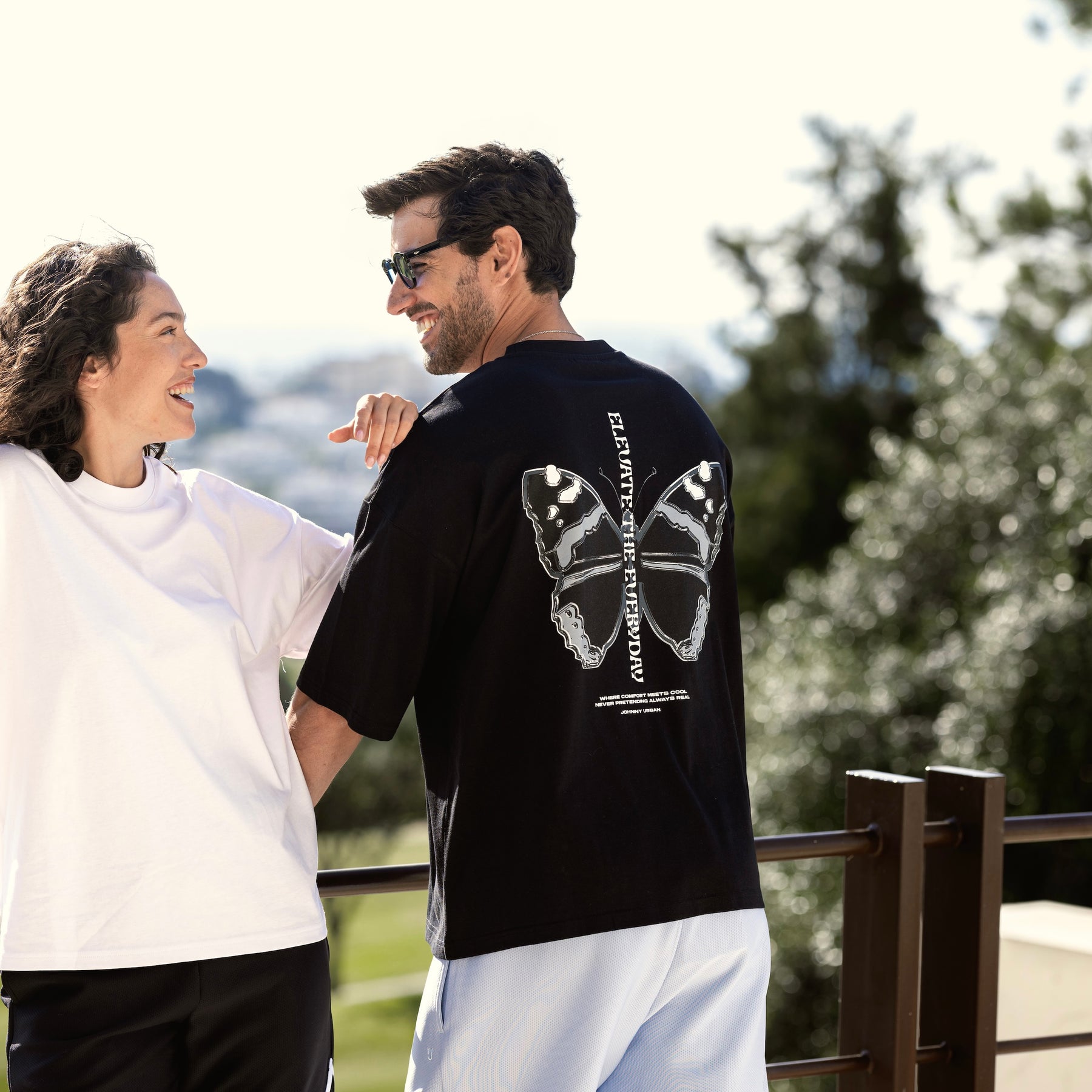 Mann trägt schwarzes Oversize-T-Shirt 'Sammy Chrome Butterfly' mit großem Schmetterlingsprint auf dem Rücken, Frau daneben lächelnd