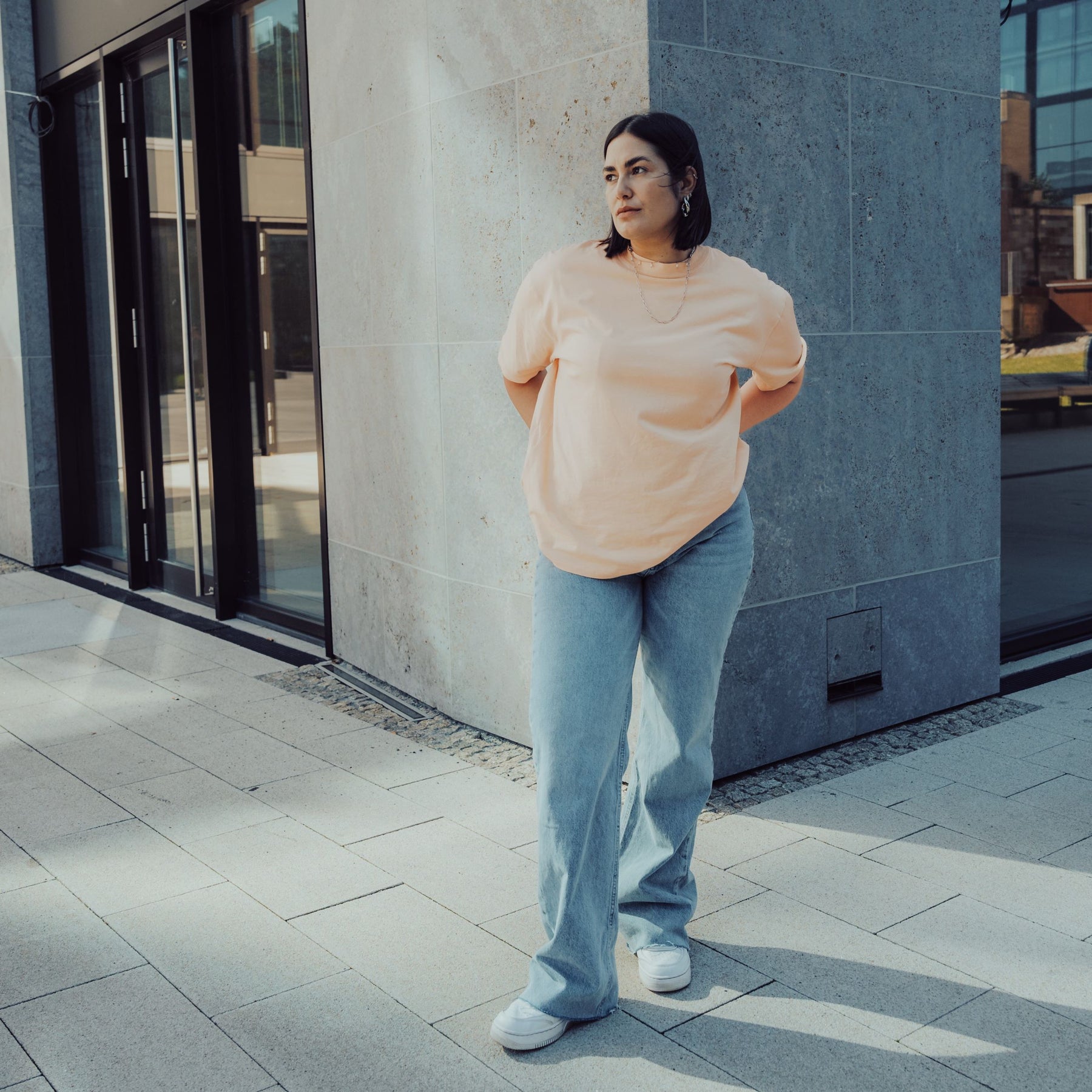 Frau trägt Oversize-T-Shirt 'Sammy' in Pfirsich-Orange und weite blaue Jeans vor moderner Fassade