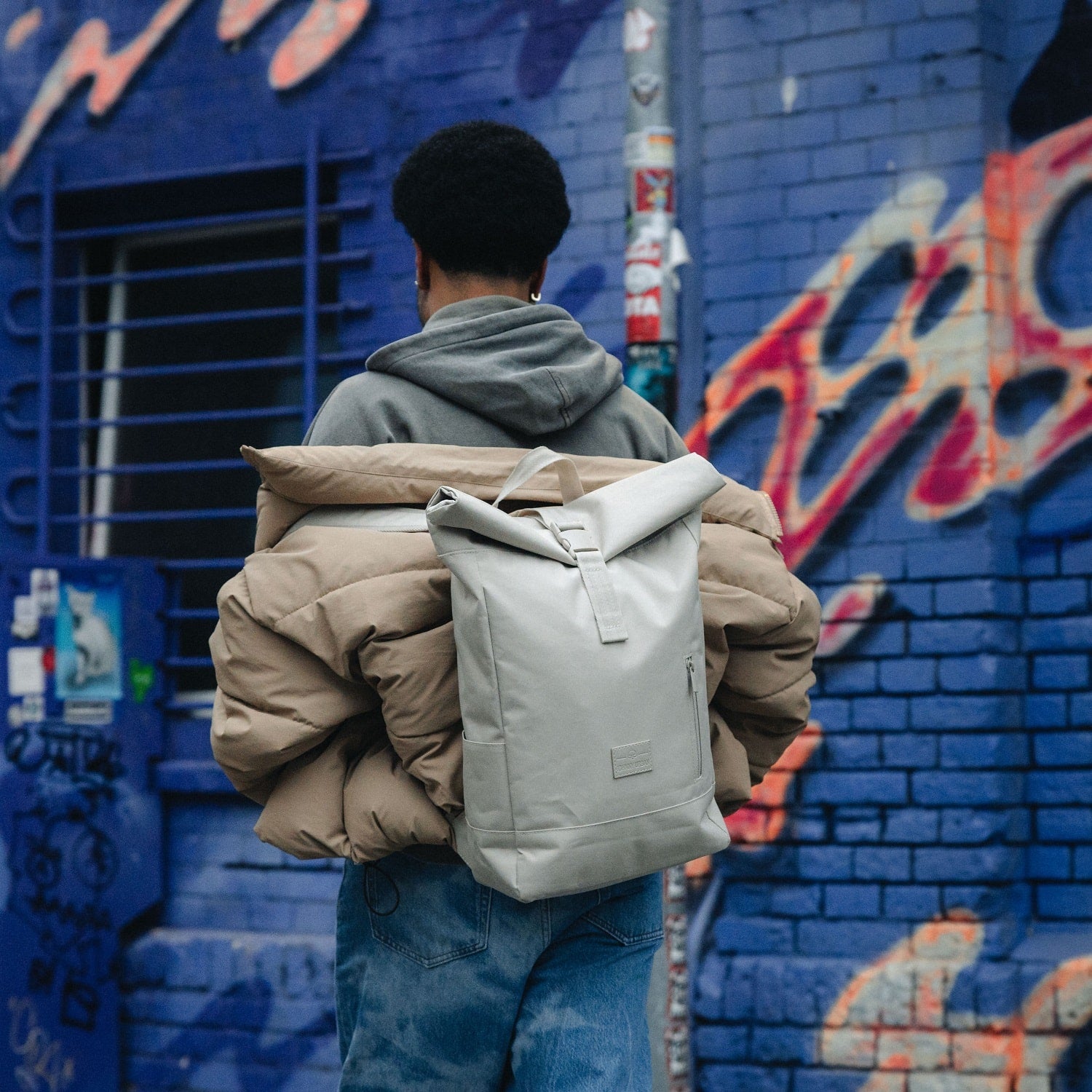 Mann trägt beigen Rolltop Rucksack 'Robin Medium' von Johnny Urban und beige Jacke vor blauem Graffiti-Hintergrund