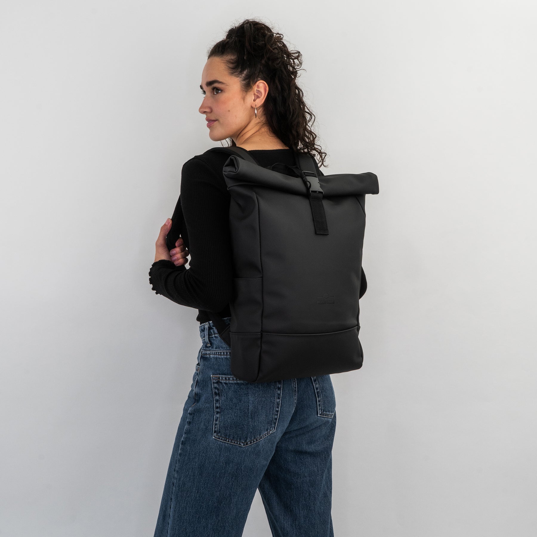 Frau mit lockigem Haar trägt schwarzen Rolltop-Rucksack 'Harvey Medium' von Johnny Urban an schwarzem Oberteil und blauen Jeans