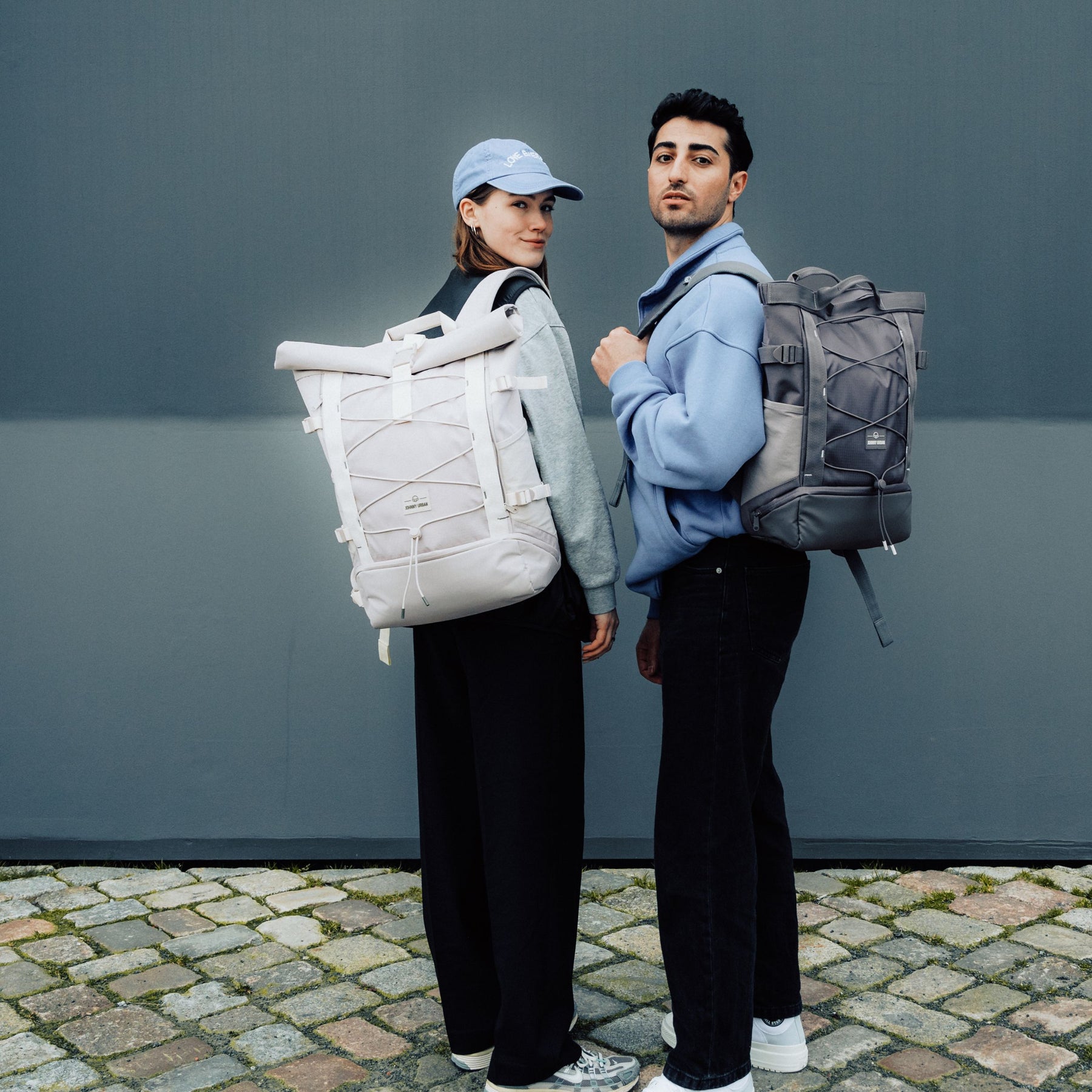 Zwei Personen tragen Rolltop Rucksäcke 'Allen Large Move', eine beige und eine graue, auf gepflastertem Boden vor grauer Wand