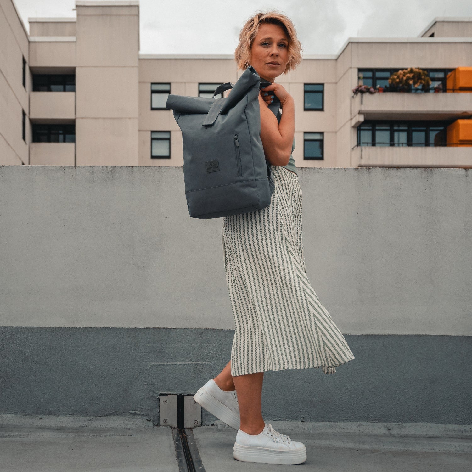 Frau trägt dunkelgrauen Rolltop Rucksack ‚Robin Medium‘, gestreifter Midirock, weiße Sneaker in urbaner Umgebung