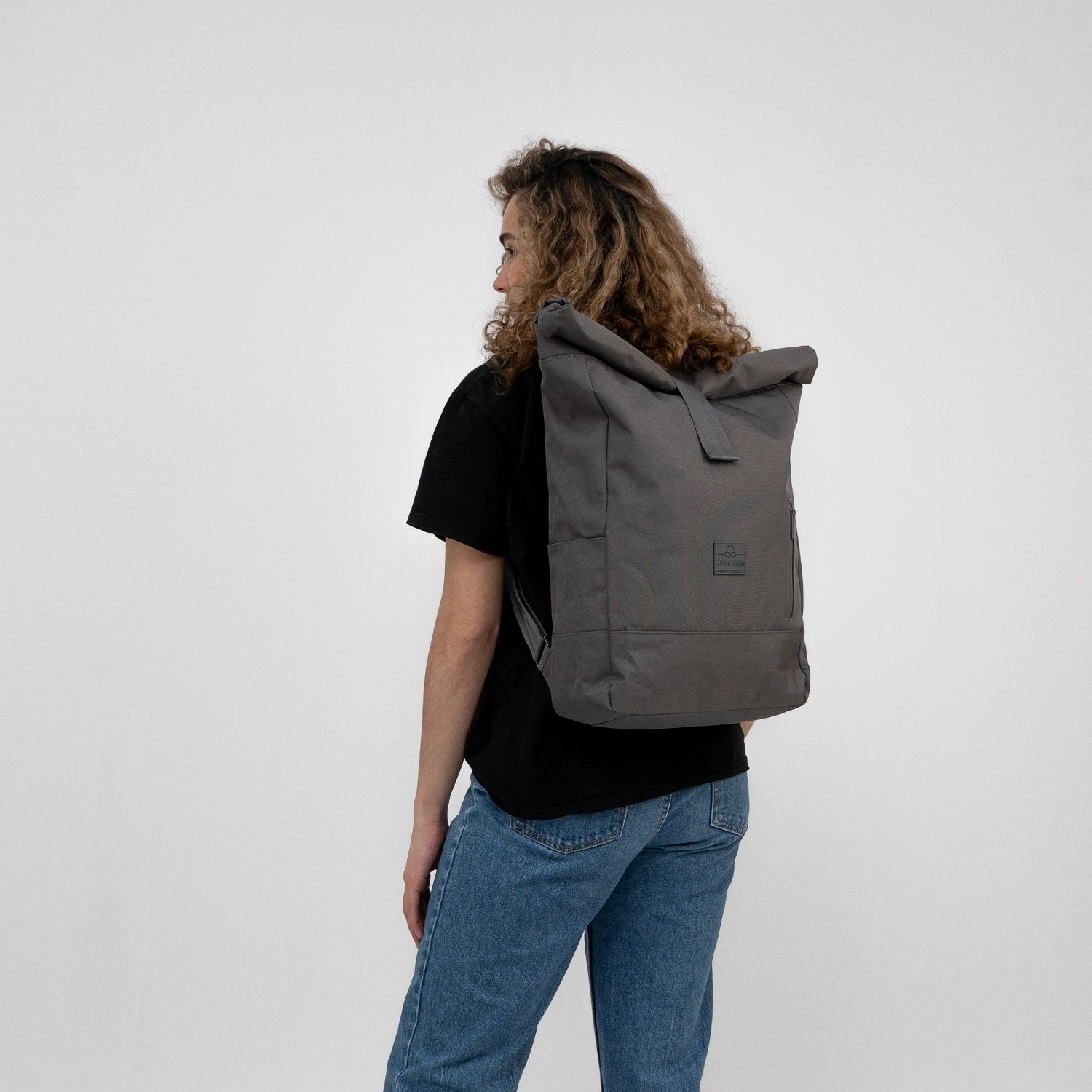 Dunkelgrauer Rolltop-Rucksack 'Robin Large' von Johnny Urban an einer Frau, seitlich stehend, mit lockigen Haaren, schwarzem T-Shirt und blauen Jeans