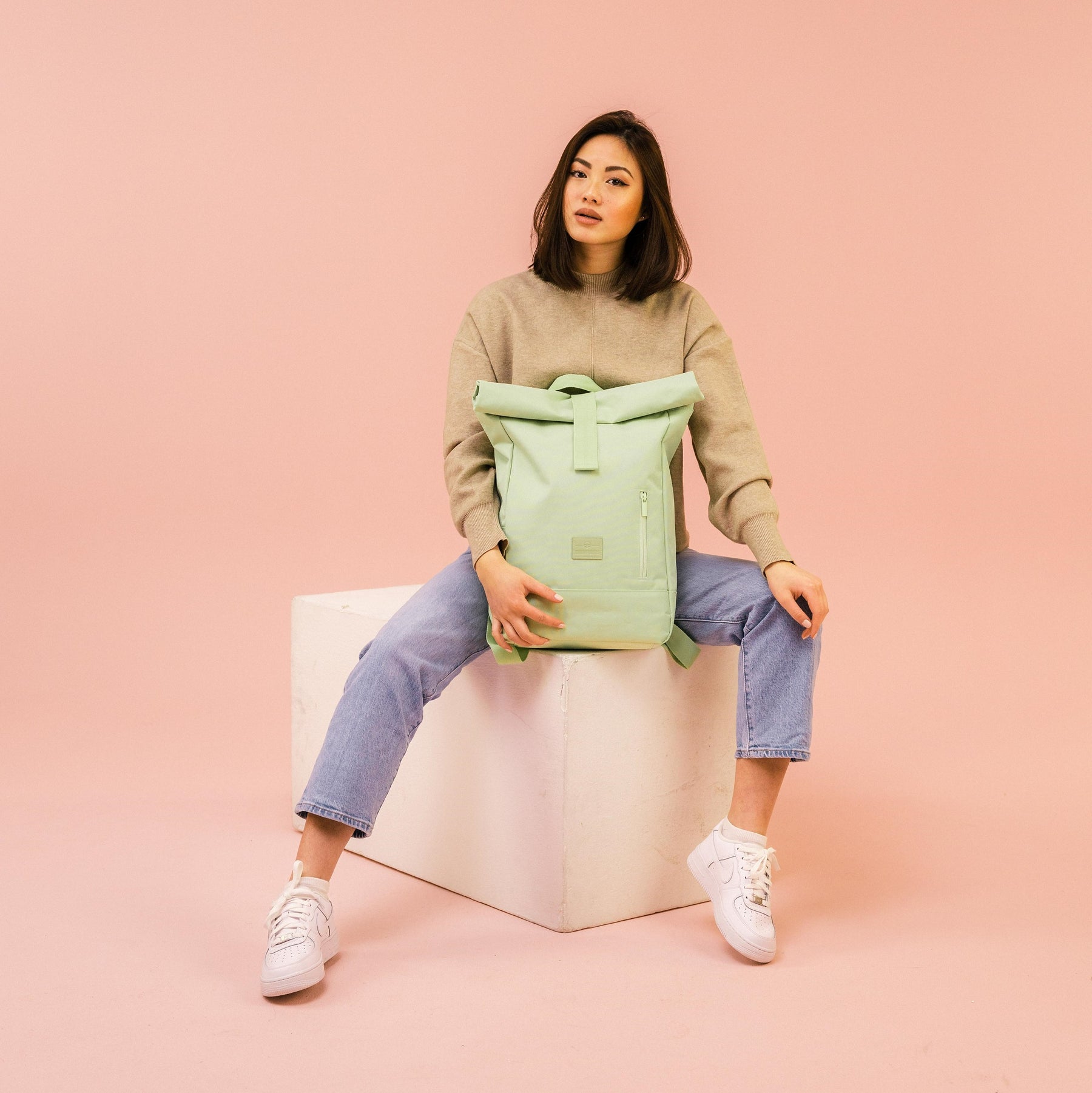 Frau sitzt auf weißem Würfel, hält limettengrünen Rolltop Rucksack Robin Medium