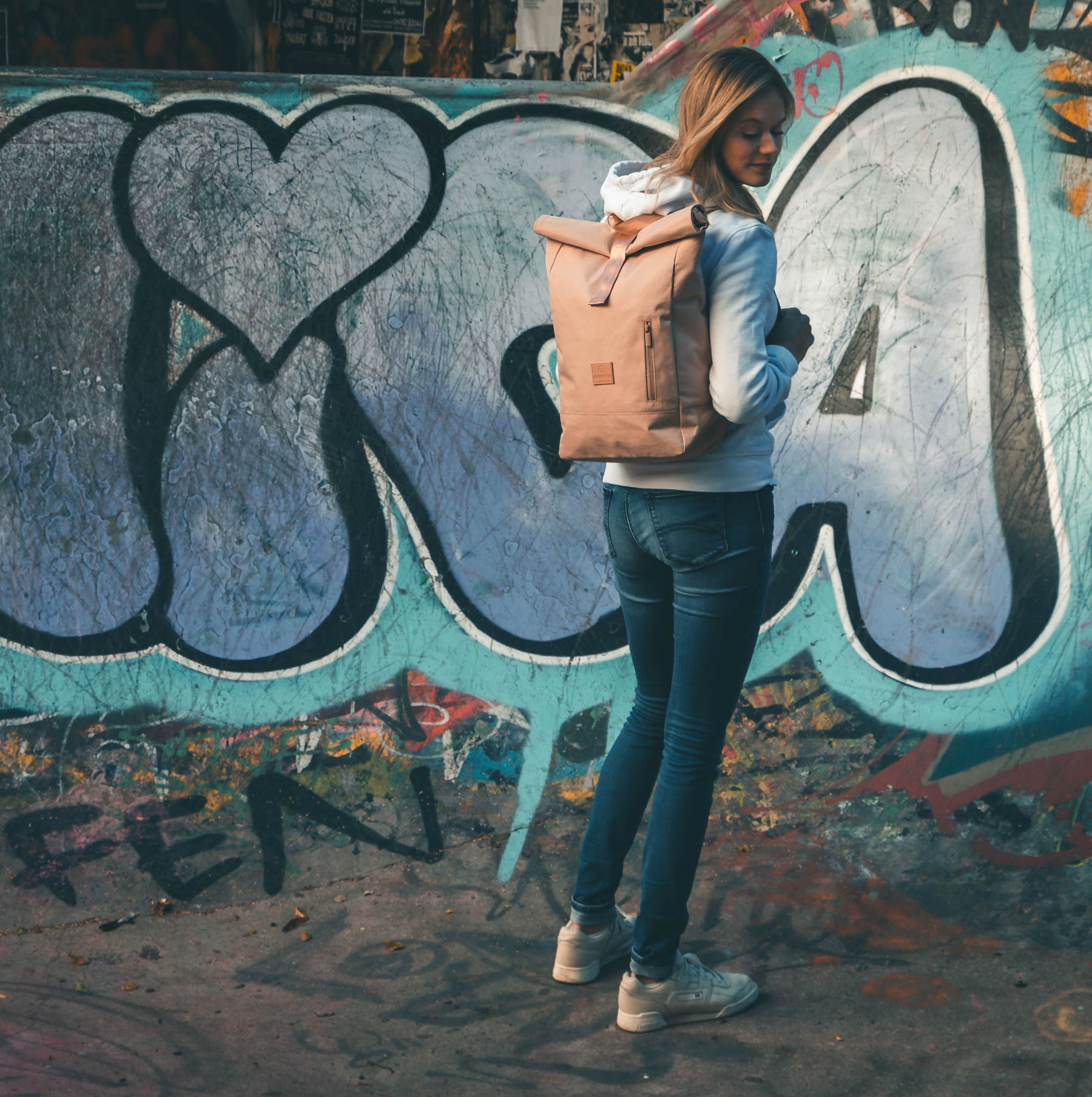 Frau mit hellrosa Rolltop-Rucksack 'Robin Medium' von Johnny Urban vor einer mit Graffiti bedeckten Wand