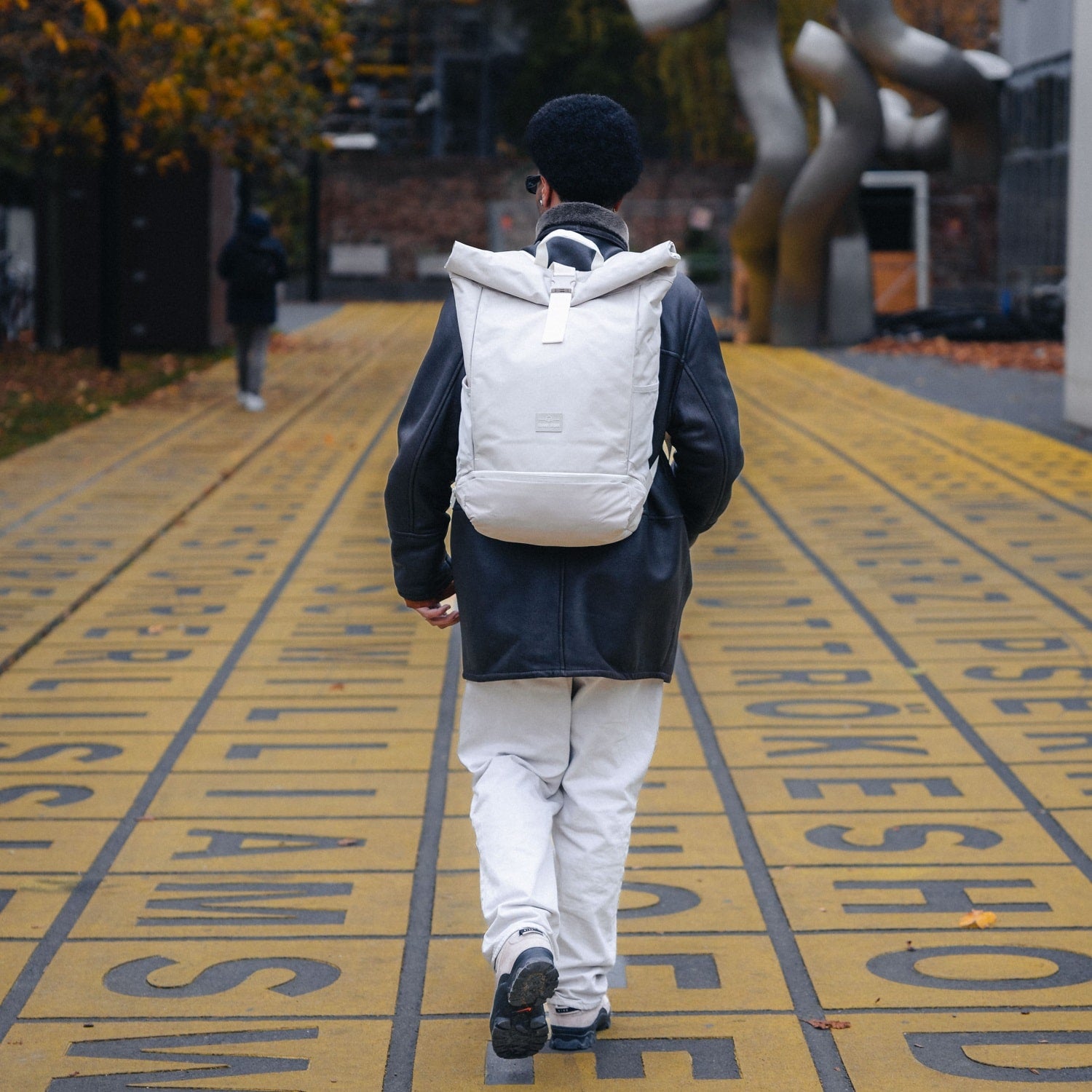Rolltop Rucksack Allen XL in Beige getragen von Mann mit schwarzer Lederjacke und weißer Hose auf gelbem Weg mit schwarzen Schriftzügen