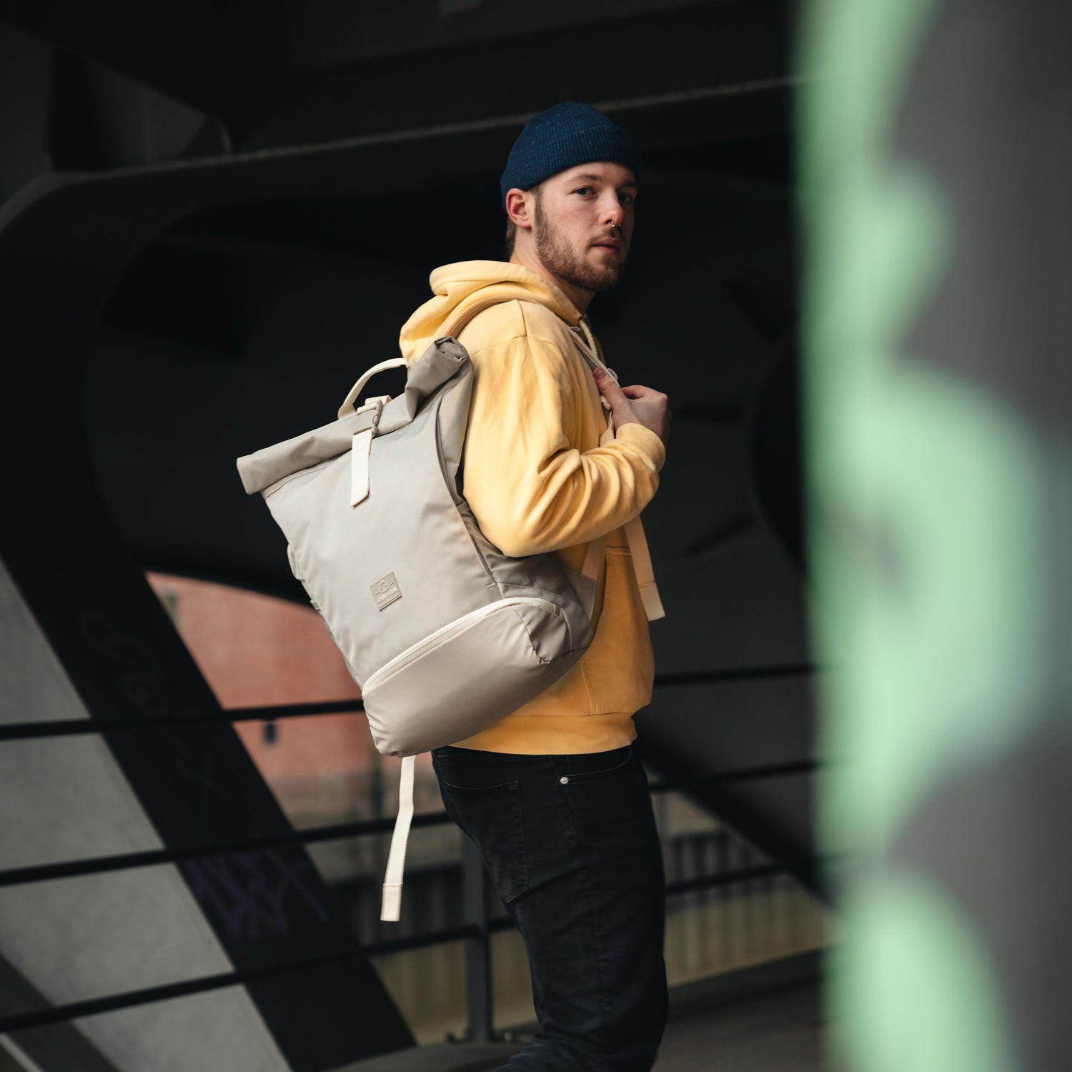 Mann mit blauem Strickmütze und gelbem Hoodie trägt beigen Rolltop Rucksack 'Allen Large' von Johnny Urban