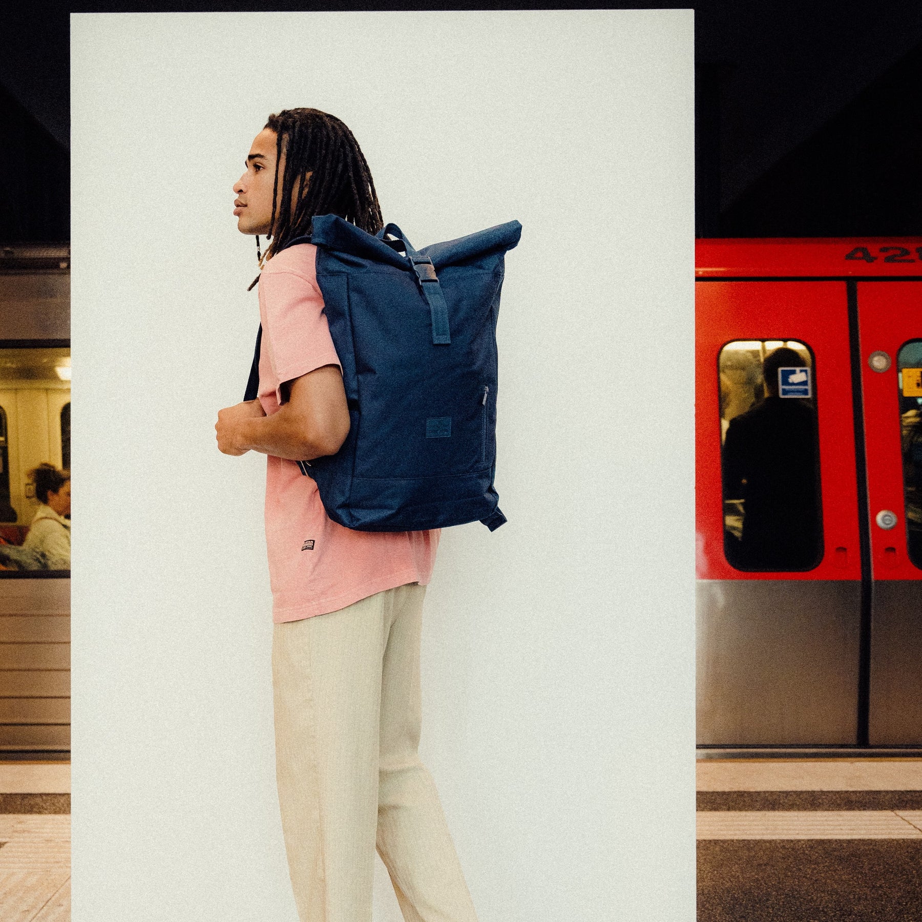 Mann mit Rolltop-Rucksack 'Robin Large' in Dunkelblau, trägt rosa T-Shirt und helle Hose an U-Bahn-Station