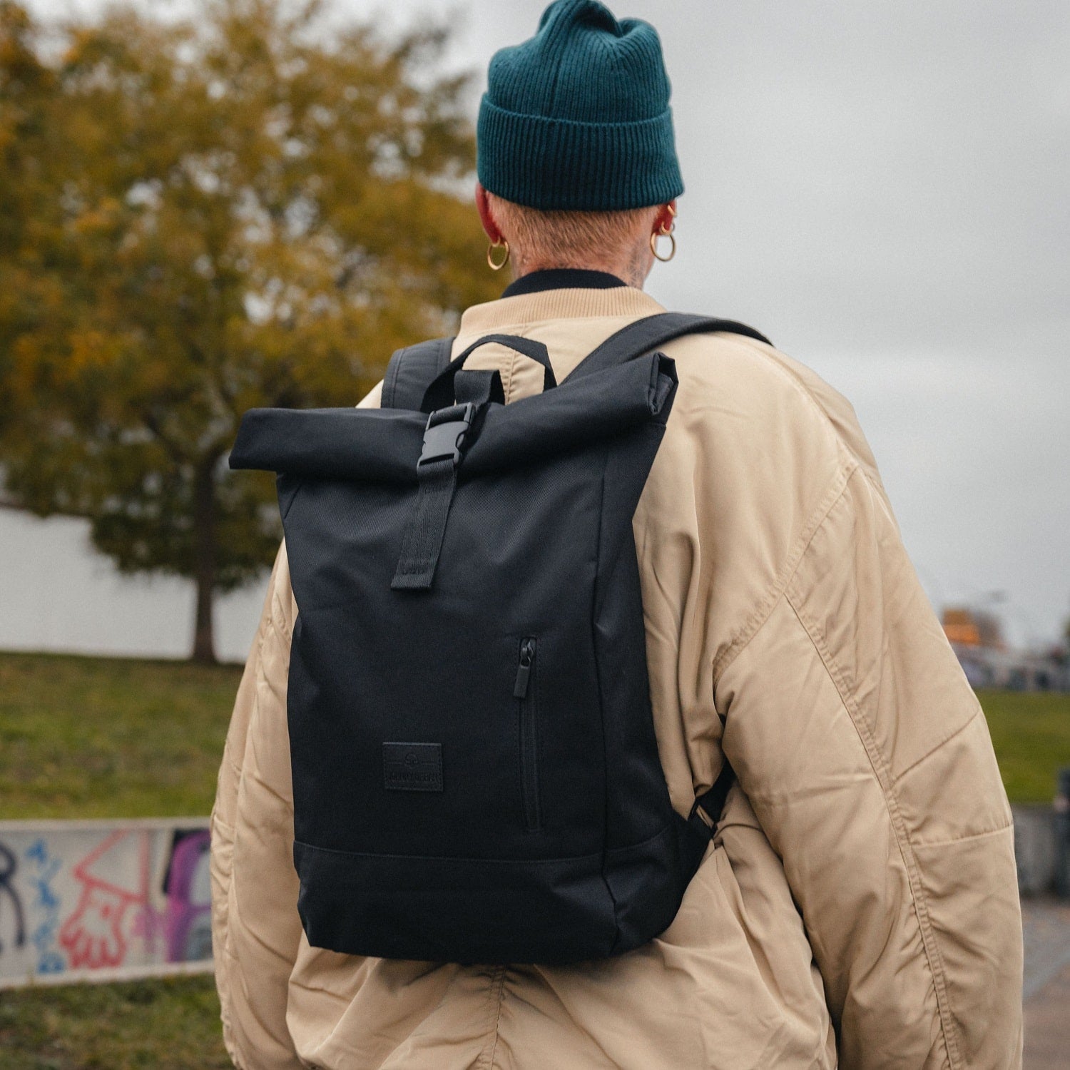 Dame trägt schwarzen Rolltop-Rucksack 'Robin Medium' von Johnny Urban mit Fronttasche, vor grauem Himmel und Herbstbaum