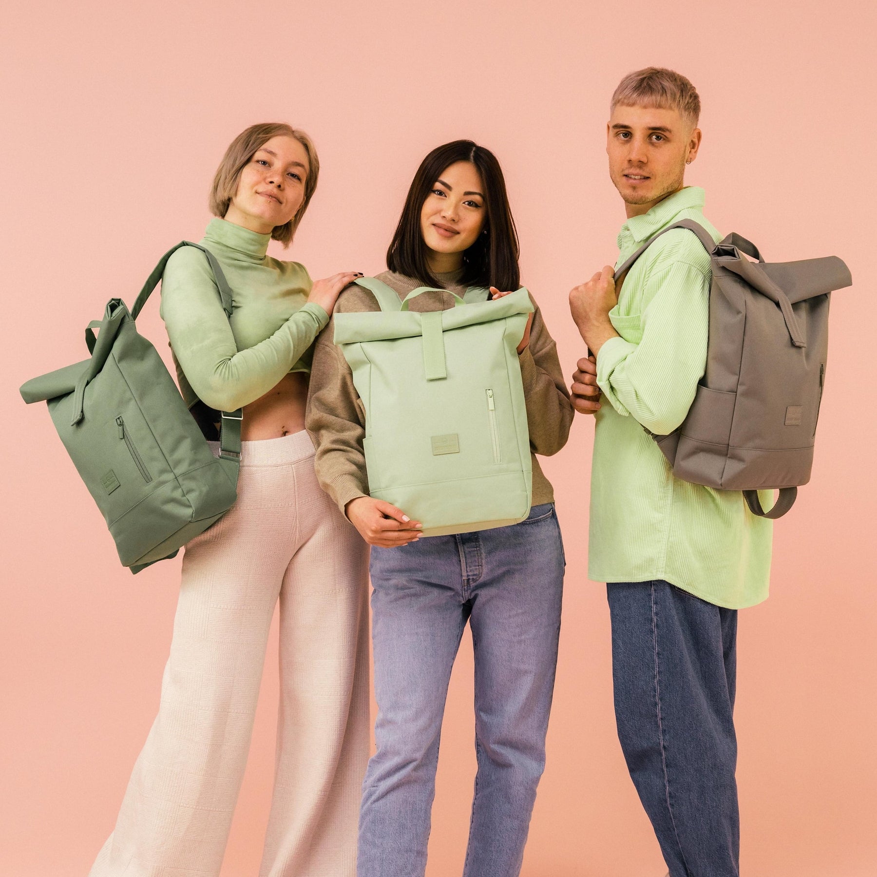 Drei Personen tragen Johnny Urban Rolltop Rucksäcke 'Robin Medium' in verschiedenen gedeckten Farben vor rosafarbenem Hintergrund