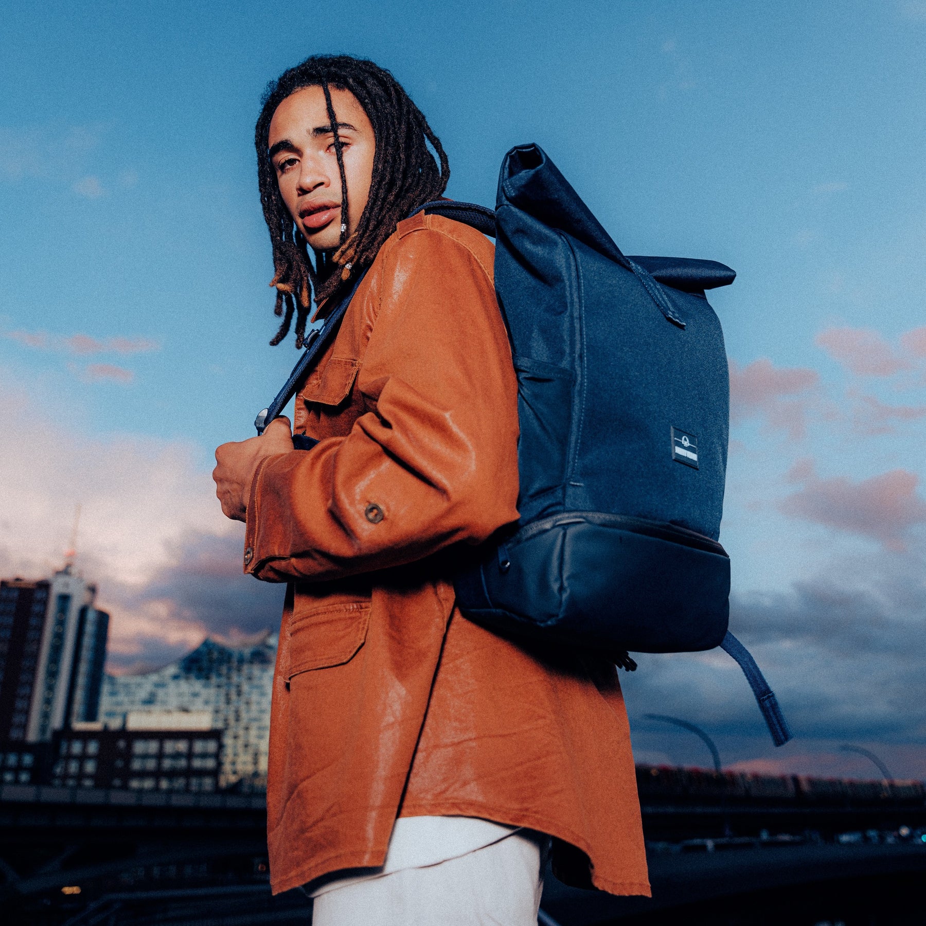 Mann trägt dunkelblauen Rolltop Rucksack 'Allen Large' von Johnny Urban und braune Jacke vor städtischer Kulisse bei Abenddämmerung