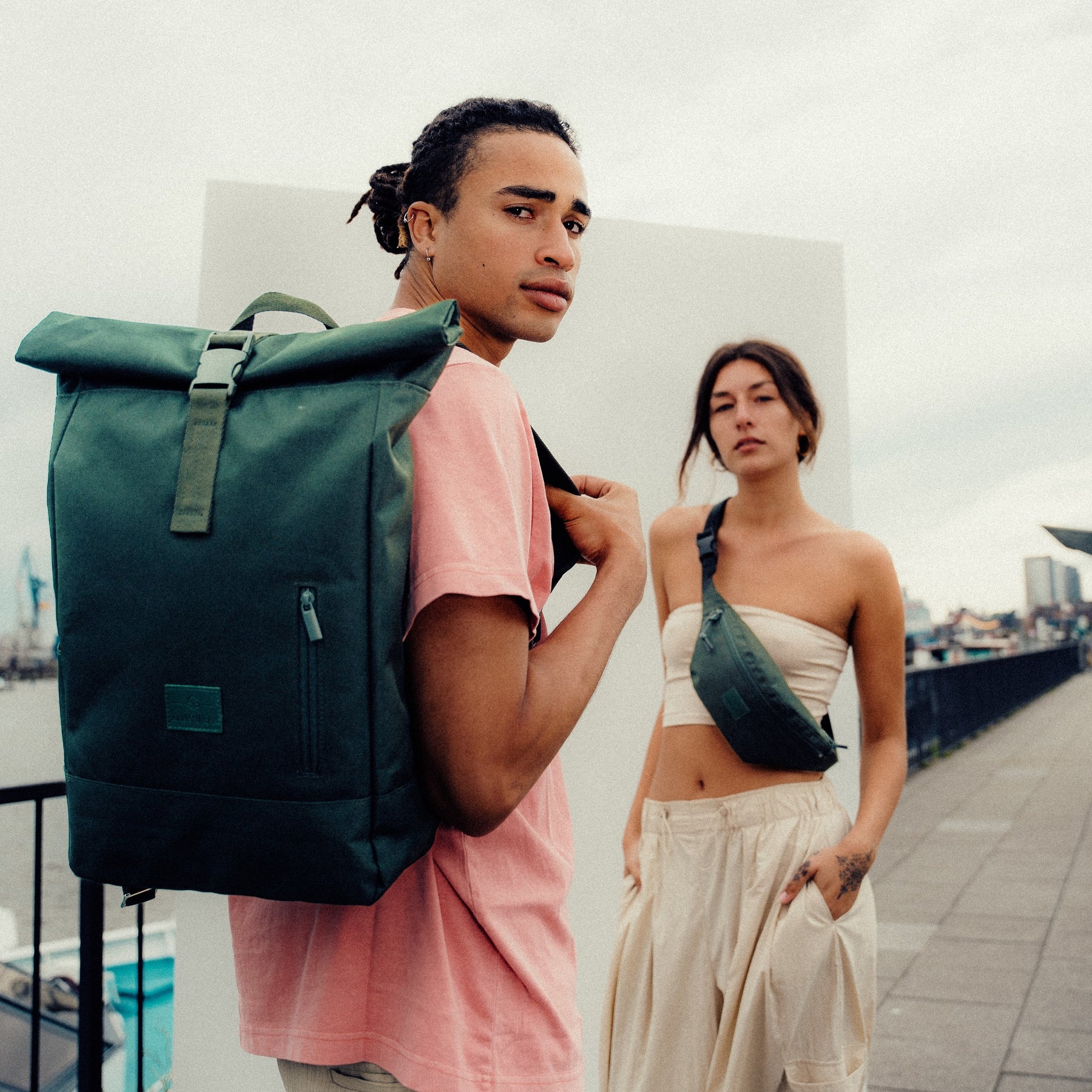 Mann mit grünem Johnny Urban Rolltop Rucksack 'Robin Large' auf dem Rücken und Frau mit grüner Umhängetasche urbanes Umfeld