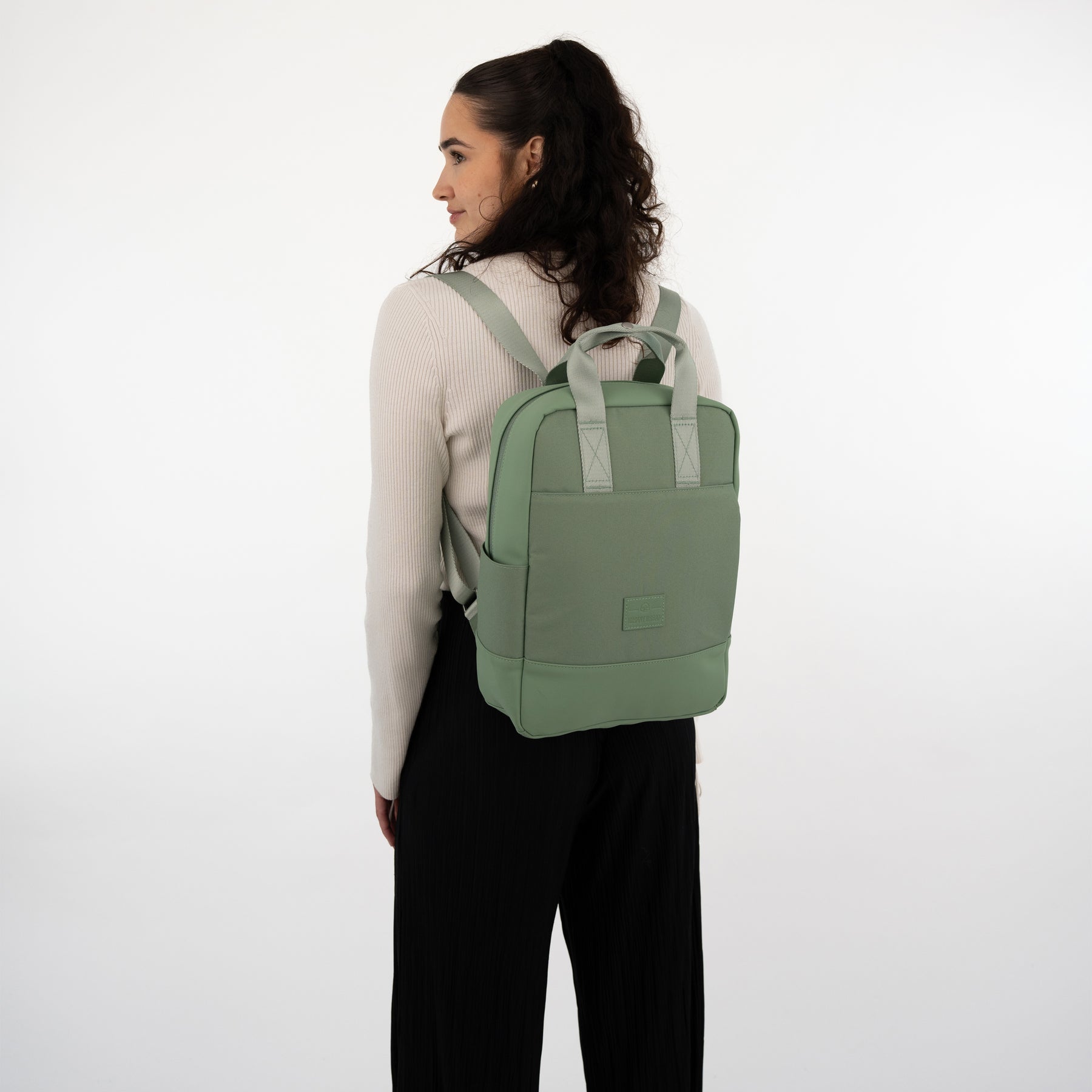 Rucksack 'Jona Medium' in Salbeigrün aus Stoff getragen von Frau mit hellem Pullover und schwarzer Hose