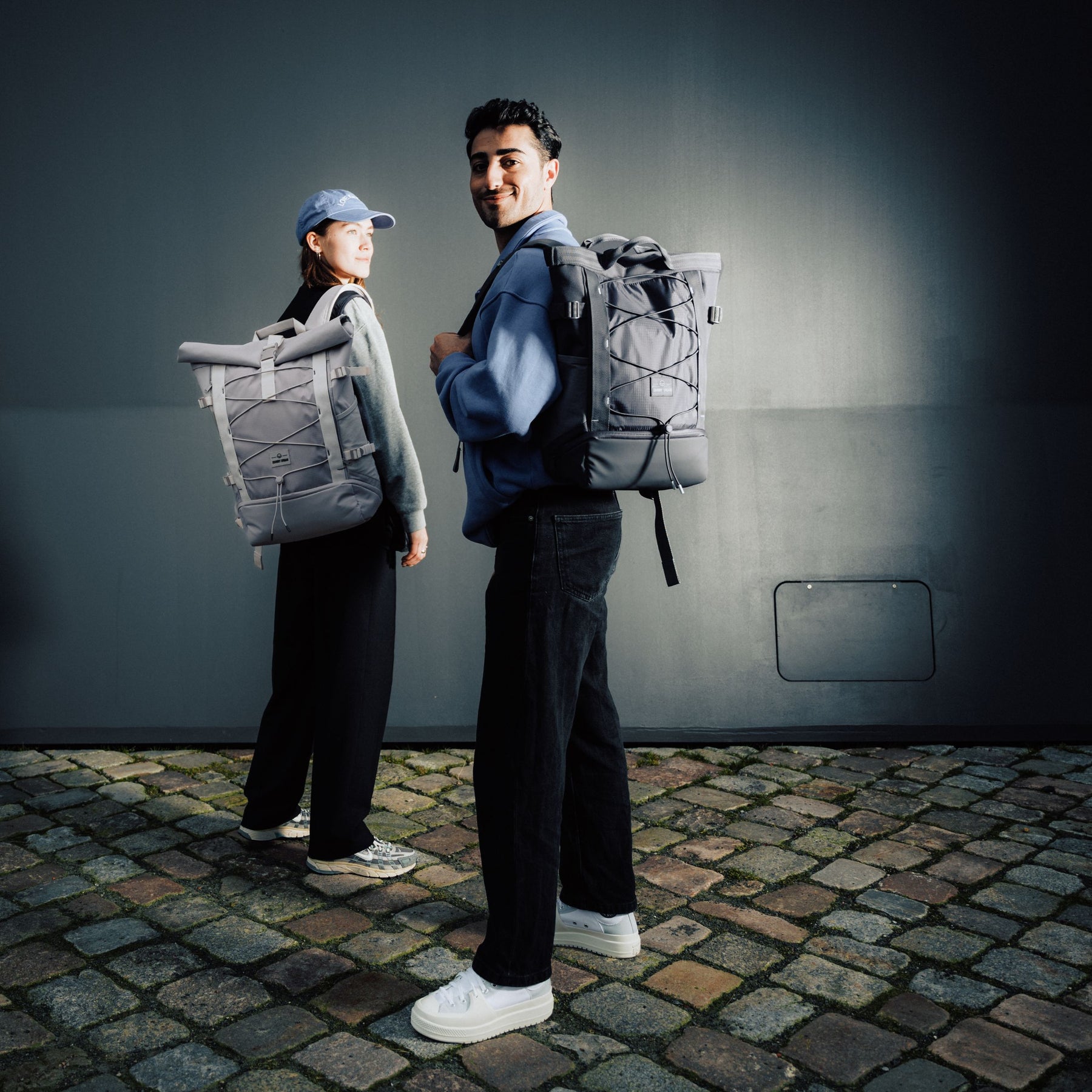 Zwei Personen mit grauen Rucksäcken 'Harper' stehend auf Kopfsteinpflaster vor dunkler Wand