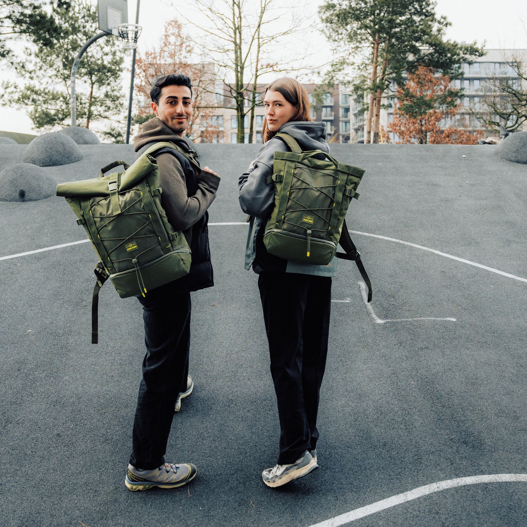 Zwei Personen, jeweils mit olivgrünem Rucksack 'Harper', auf einem urbanen Basketballplatz