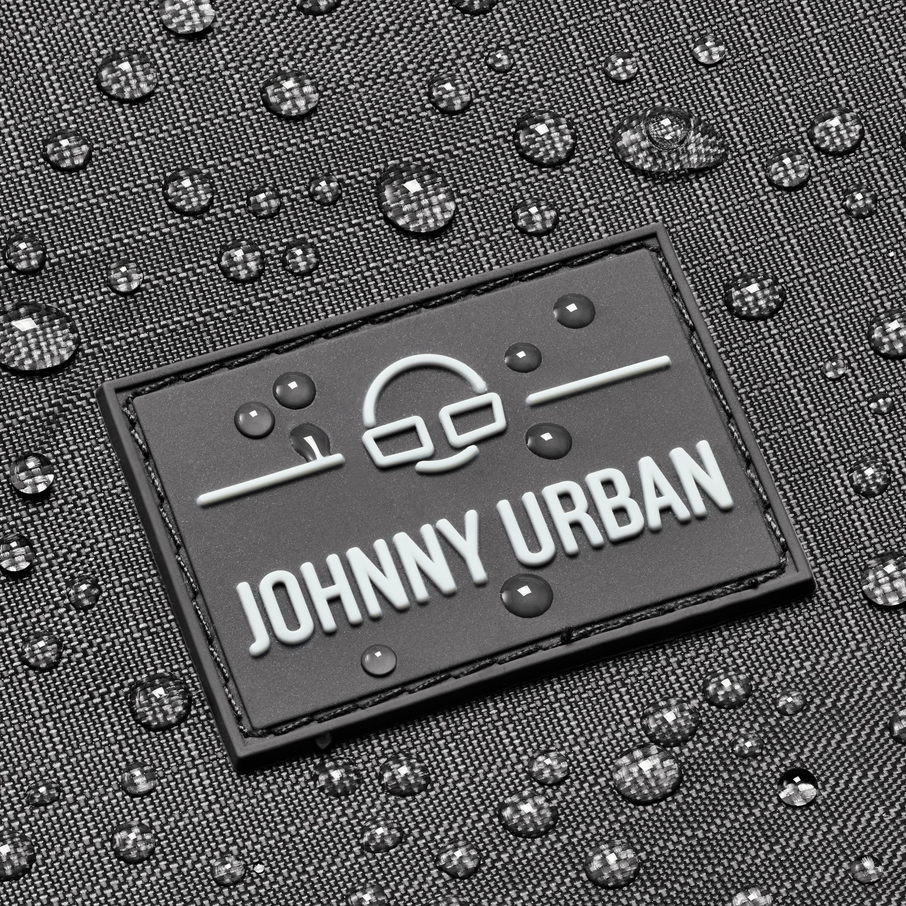 Nahaufnahme des Johnny Urban Logos auf grauem, wasserabweisendem Harper Rucksack mit Wasserperlen