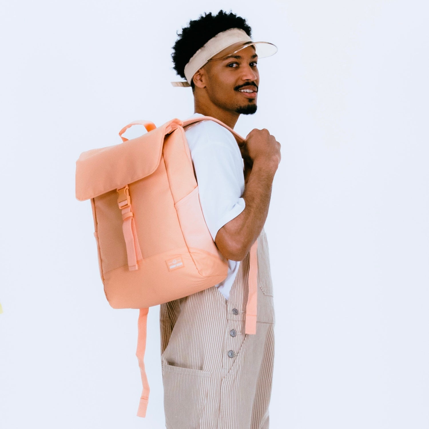 Mann trägt Rucksack "Mika" in Pfirsichfarbe, weißes T-Shirt, gestreifte Overalls und Stoff-Visier