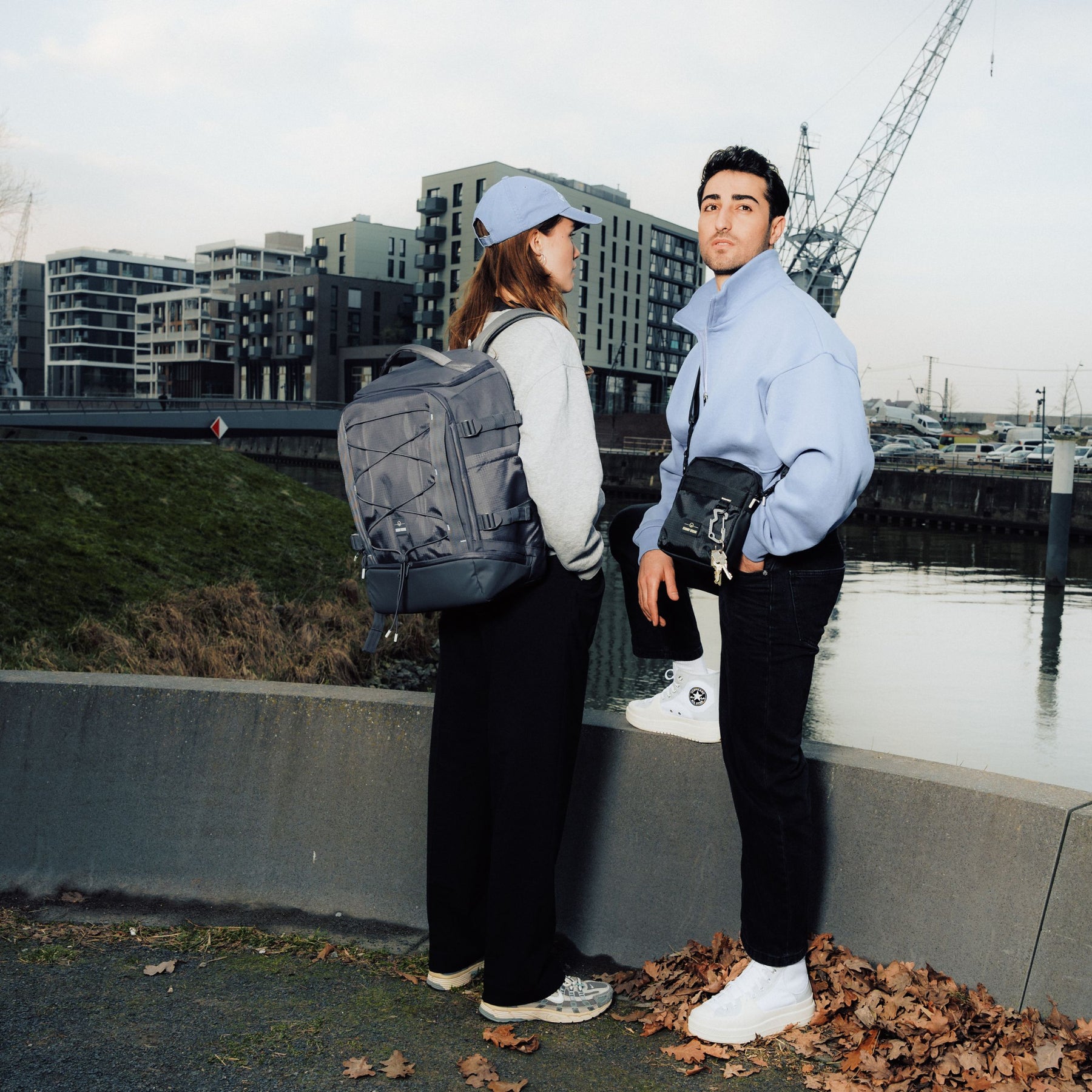 Frau mit grauem Rucksack 'Jack' und Baseballkappe, Mann mit schwarzer Umhängetasche, städtische Hafenkulisse