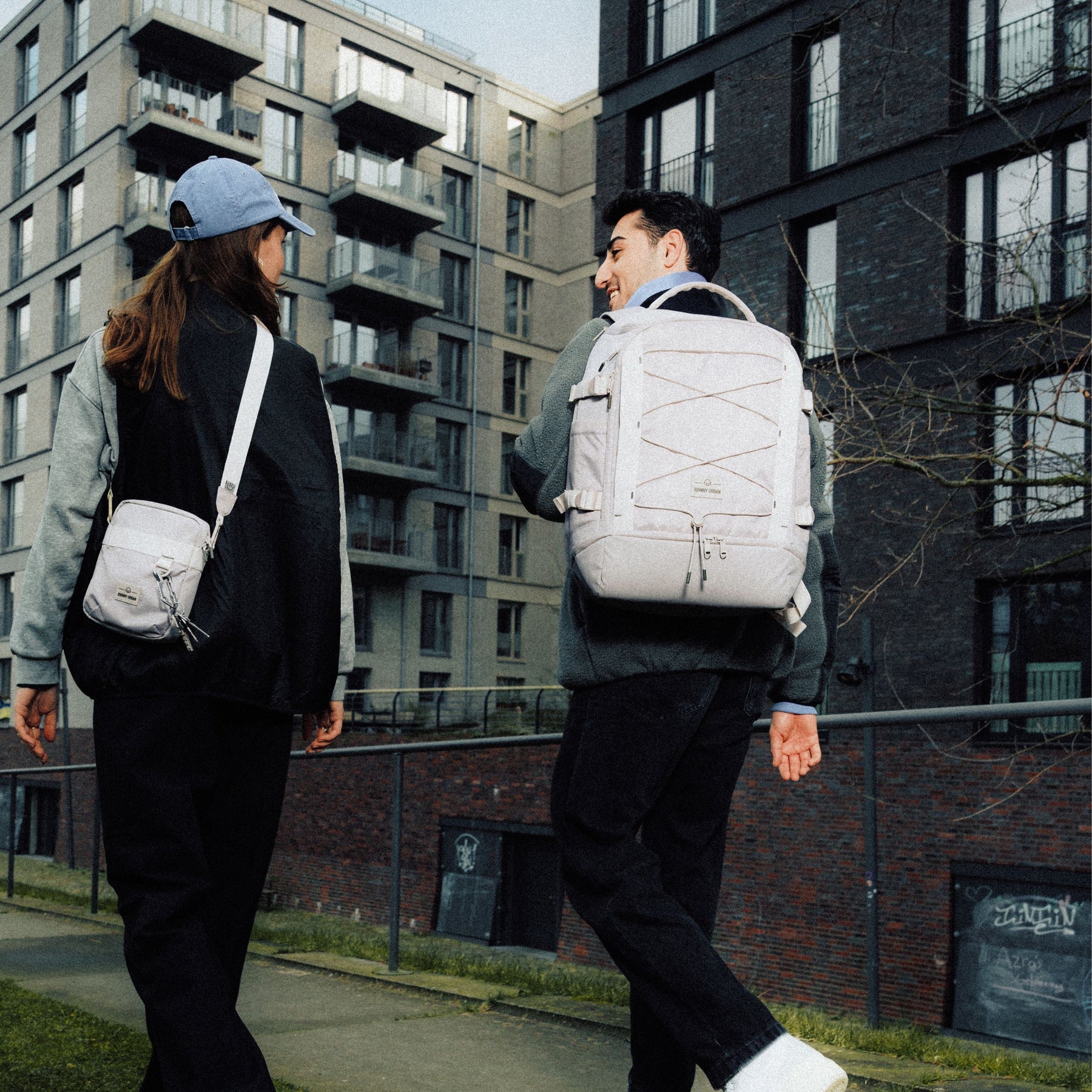 Zwei Personen gehen an einem urbanen Wohnhaus vorbei, eine trägt den beige Rucksack ‚Jack‘ von Johnny Urban
