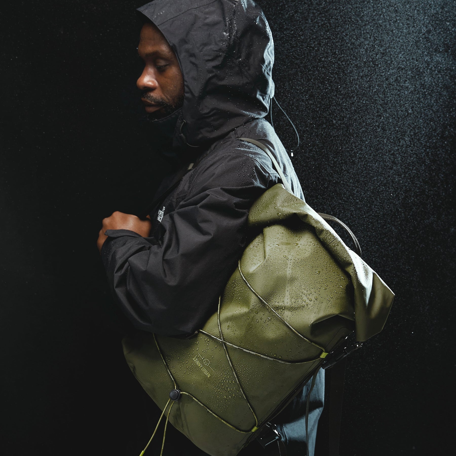 Mann in schwarzer Regenjacke trägt olivefarbenen rolltop Rucksack 'Myles' von Johnny Urban im Regen