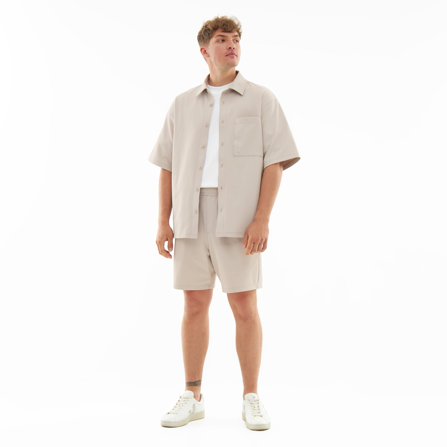 Männliches Modell trägt Sandfarbene Shorts und Kurzarm-Hemd aus dem Set 'Mason' mit weißen Turnschuhen