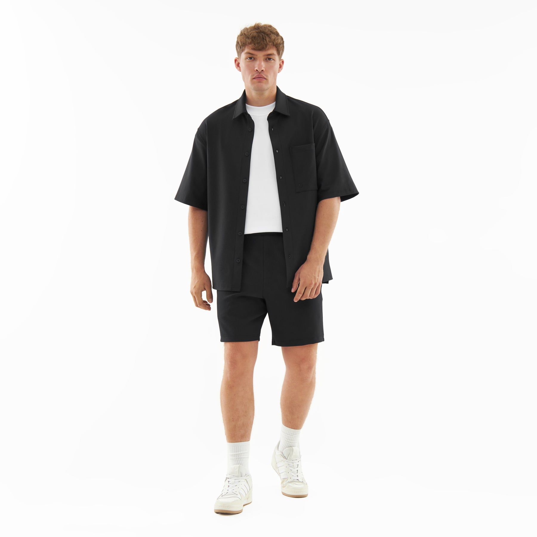 Männliches Modell trägt schwarze Shorts 'Mason', schwarze kurzärmelige Hemdjacke, weißes T-Shirt und weiße Sneakers