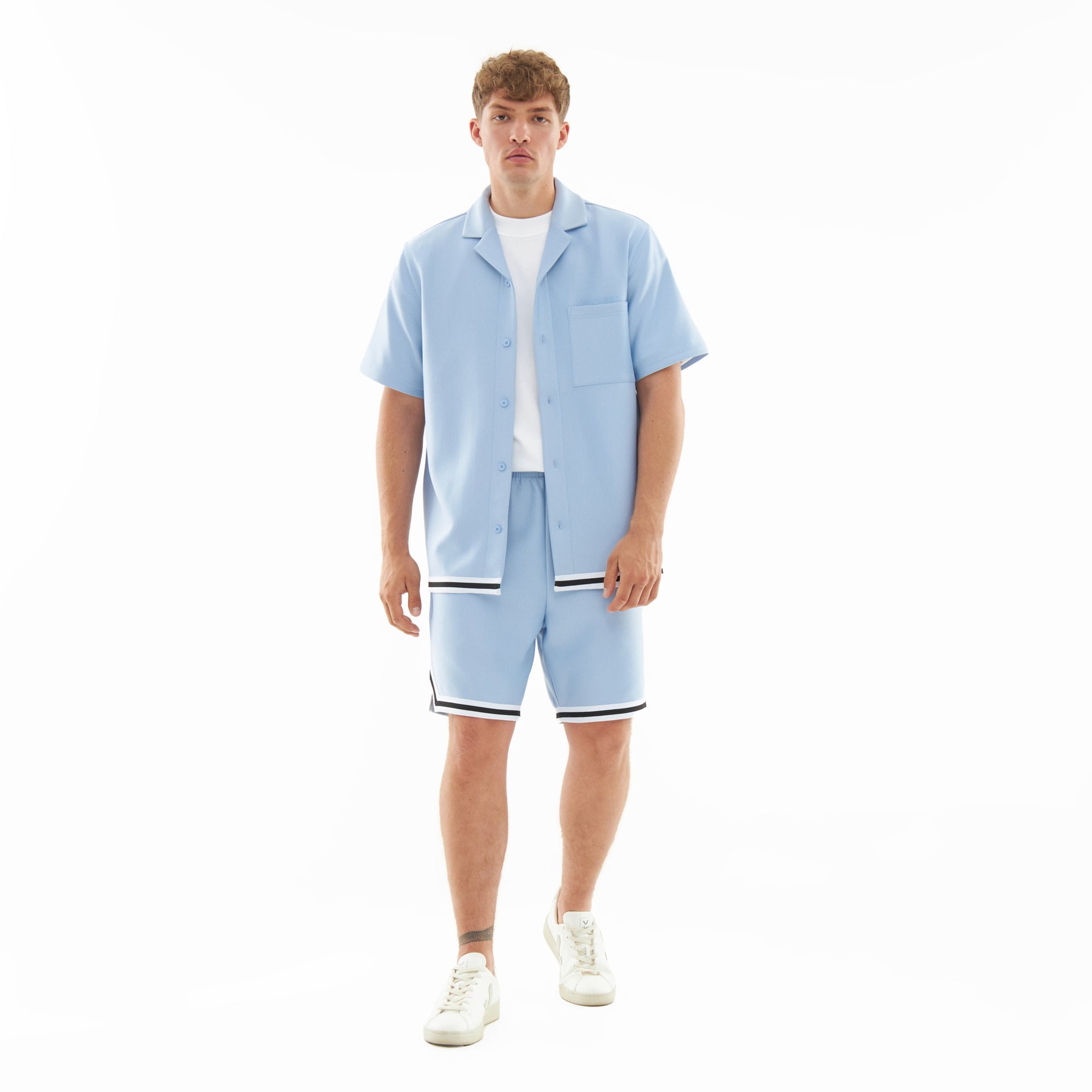 Mann trägt hellblaue Shorts 'Parker' mit kontrastierenden Streifen an Saum, kombiniert mit kurzärmligem Hemd und weißen Sneakern