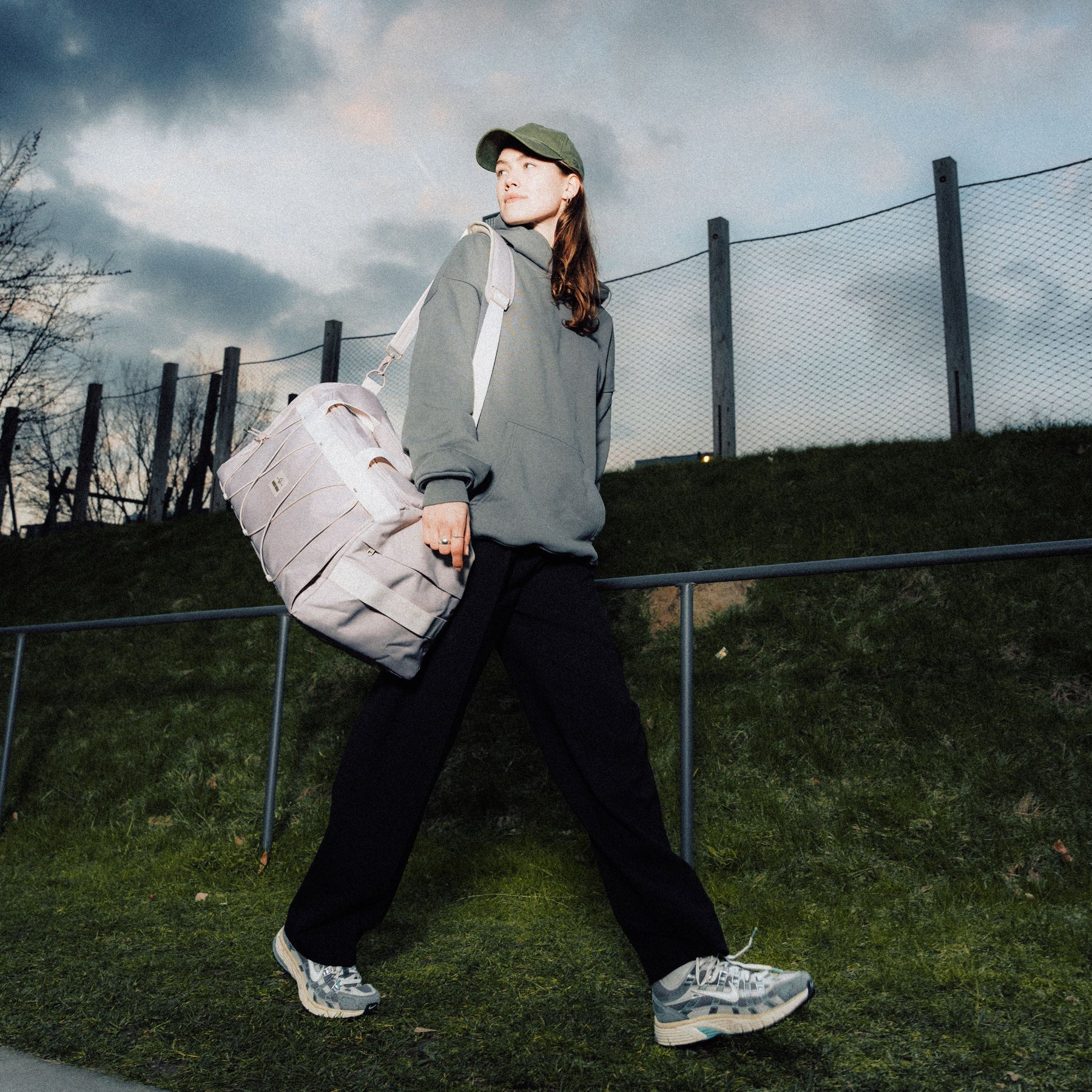 Frau trägt beige Duffle Bag "Shawn Medium" über der Schulter, trägt grünen Pullover, schwarze Hose und Baseballcap