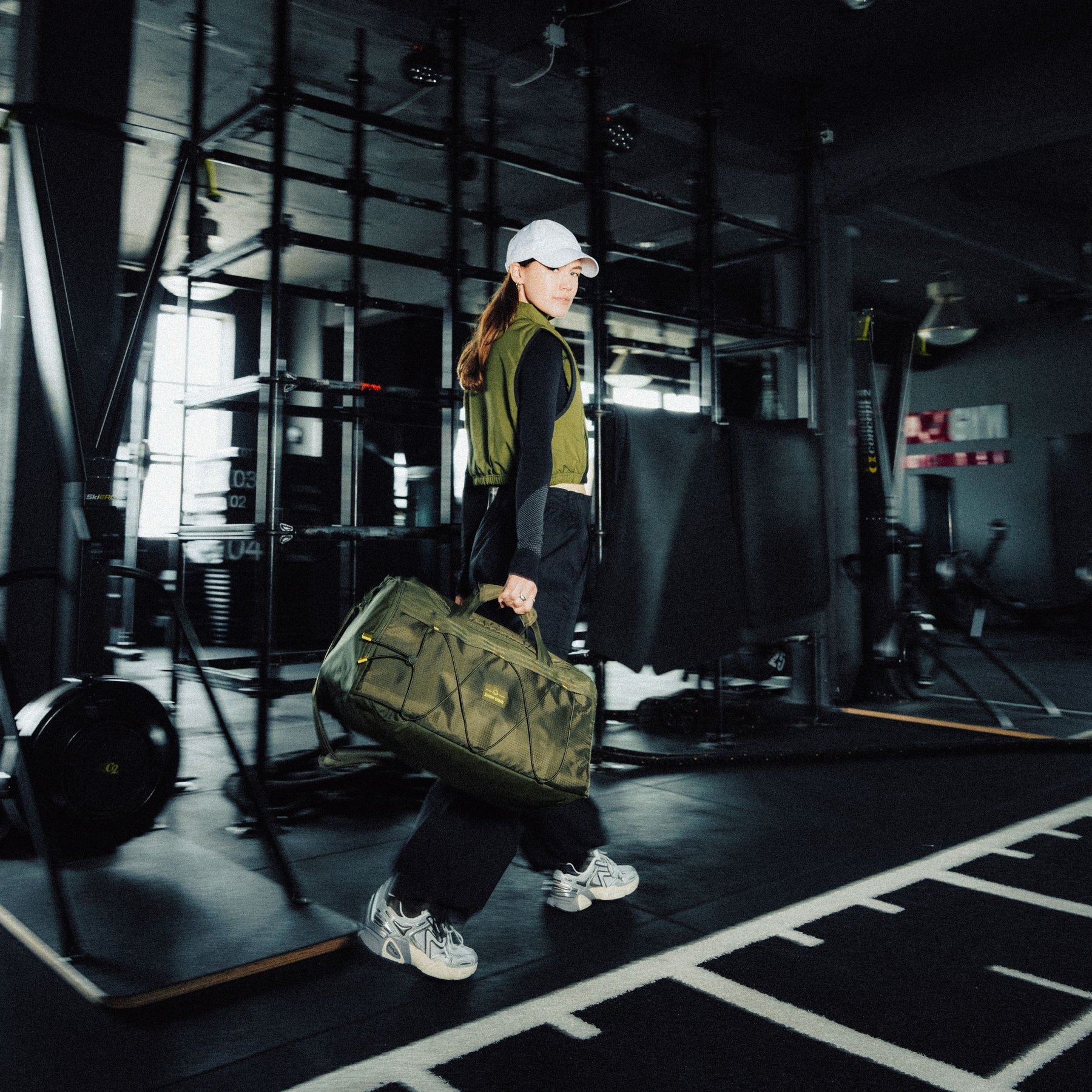 Frau in schwarzer Kleidung und olivfarbener Weste trägt Duffle Bag 'Shawn Medium' in Olivgrün im Fitnessstudio