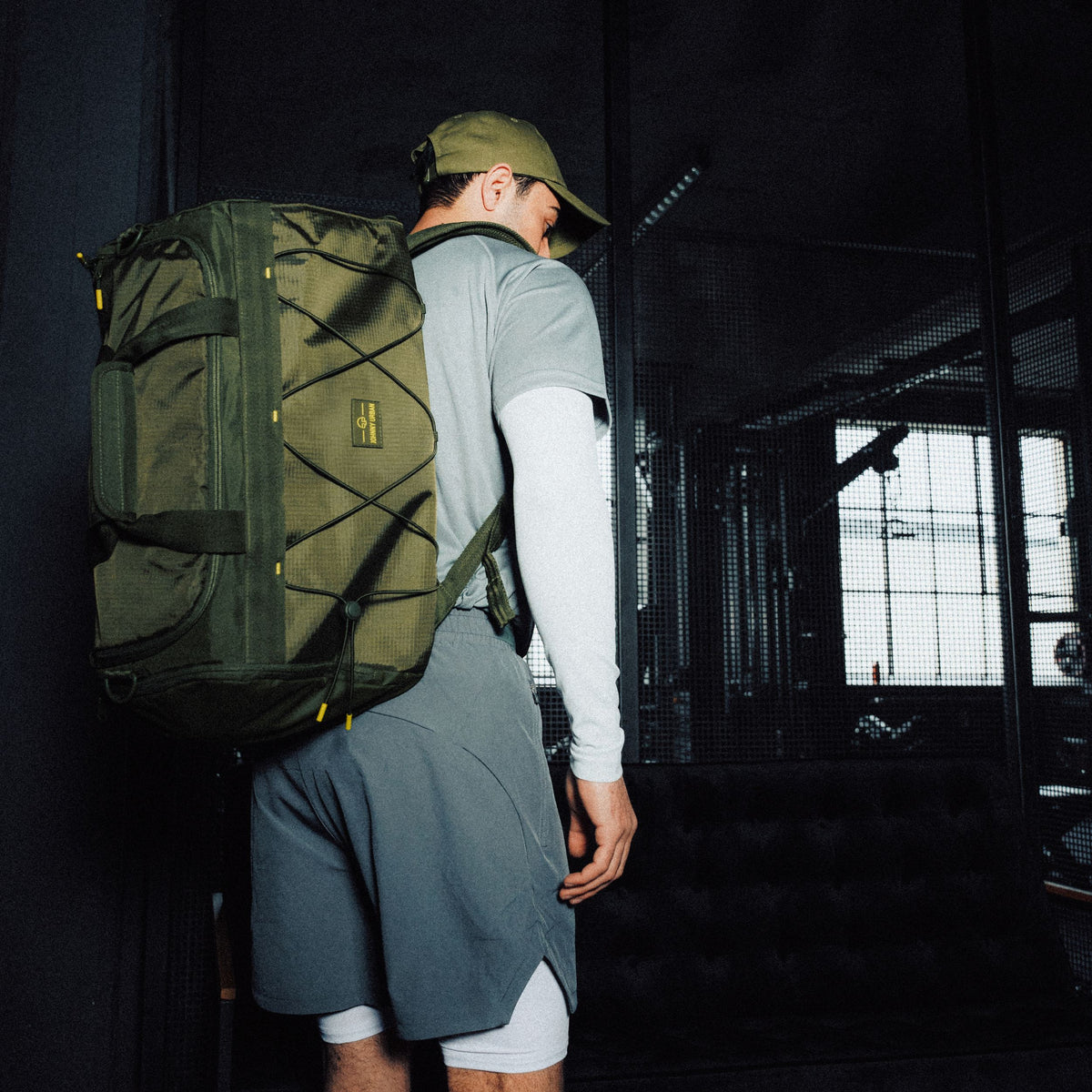 Mann mit olivgrüner Johnny Urban Duffle Bag 'Shawn Medium' im sportlichen Outfit aus grauem Shirt, Shorts und Cap