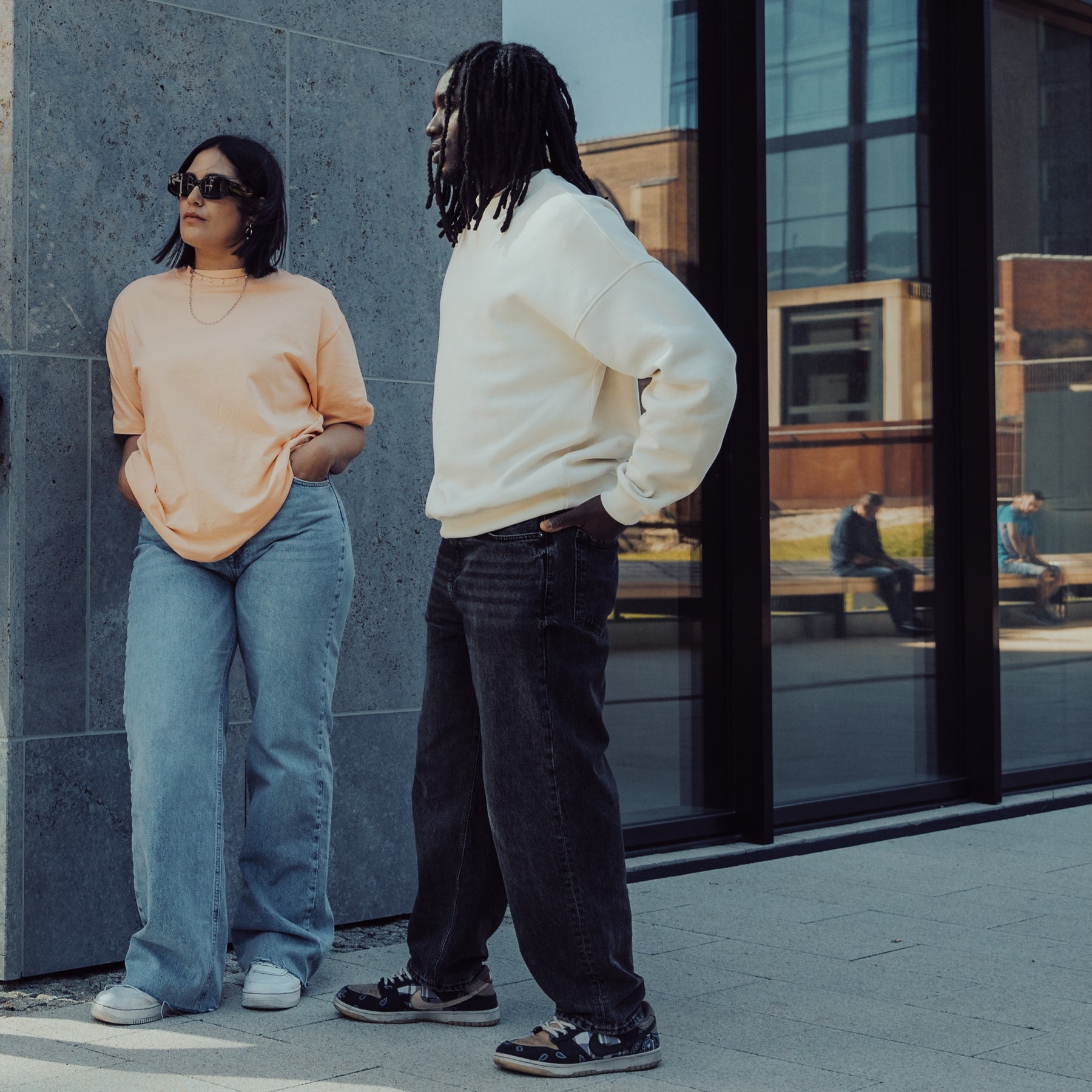 Zwei Personen vor modernem Gebäude, eine trägt weißen Oversized Crewneck Pullover 'Carter', die andere ein beiges T-Shirt