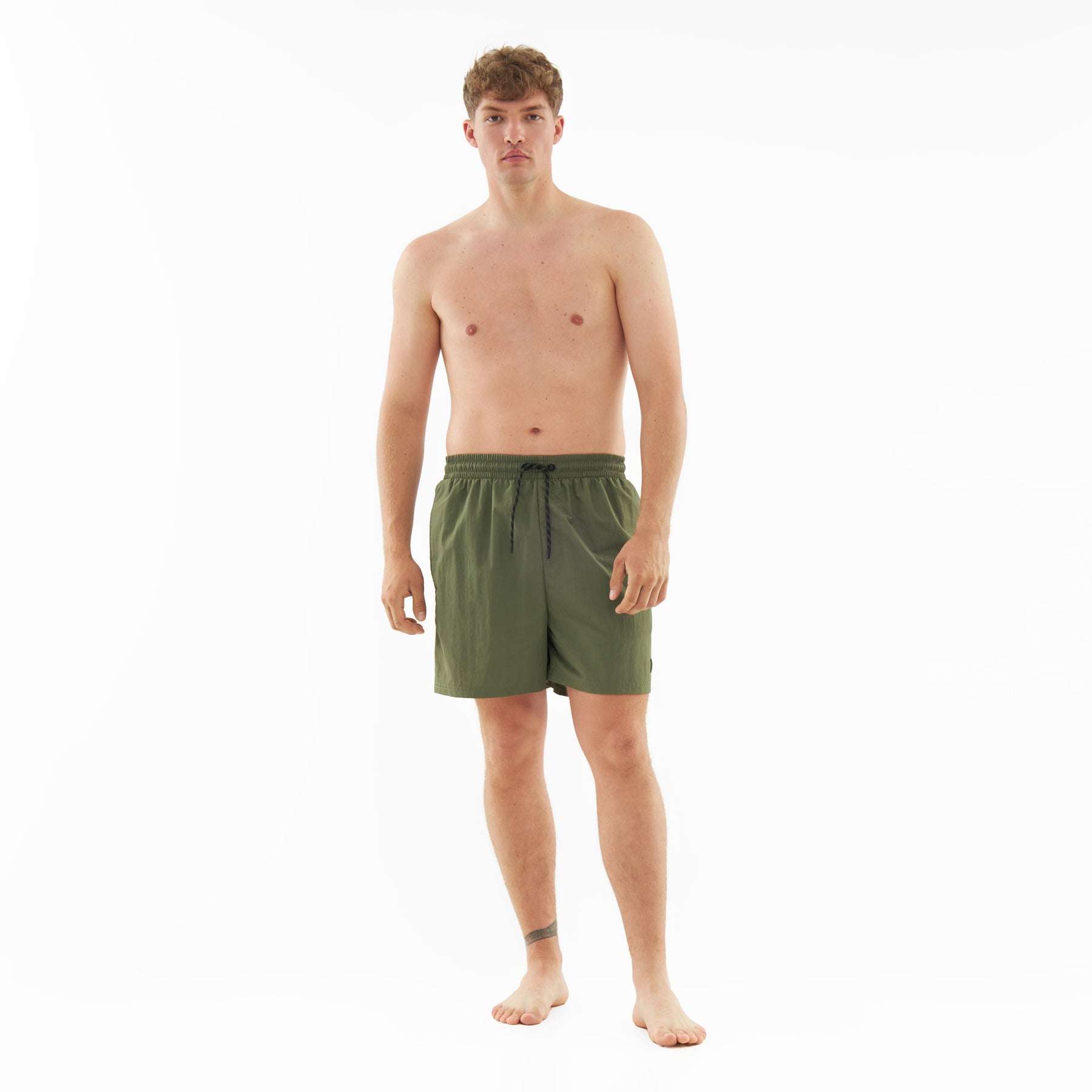 Mann trägt olivgrüne Badehose 'Jay' mit Kordelzug, steht barfuß und ohne Oberteil