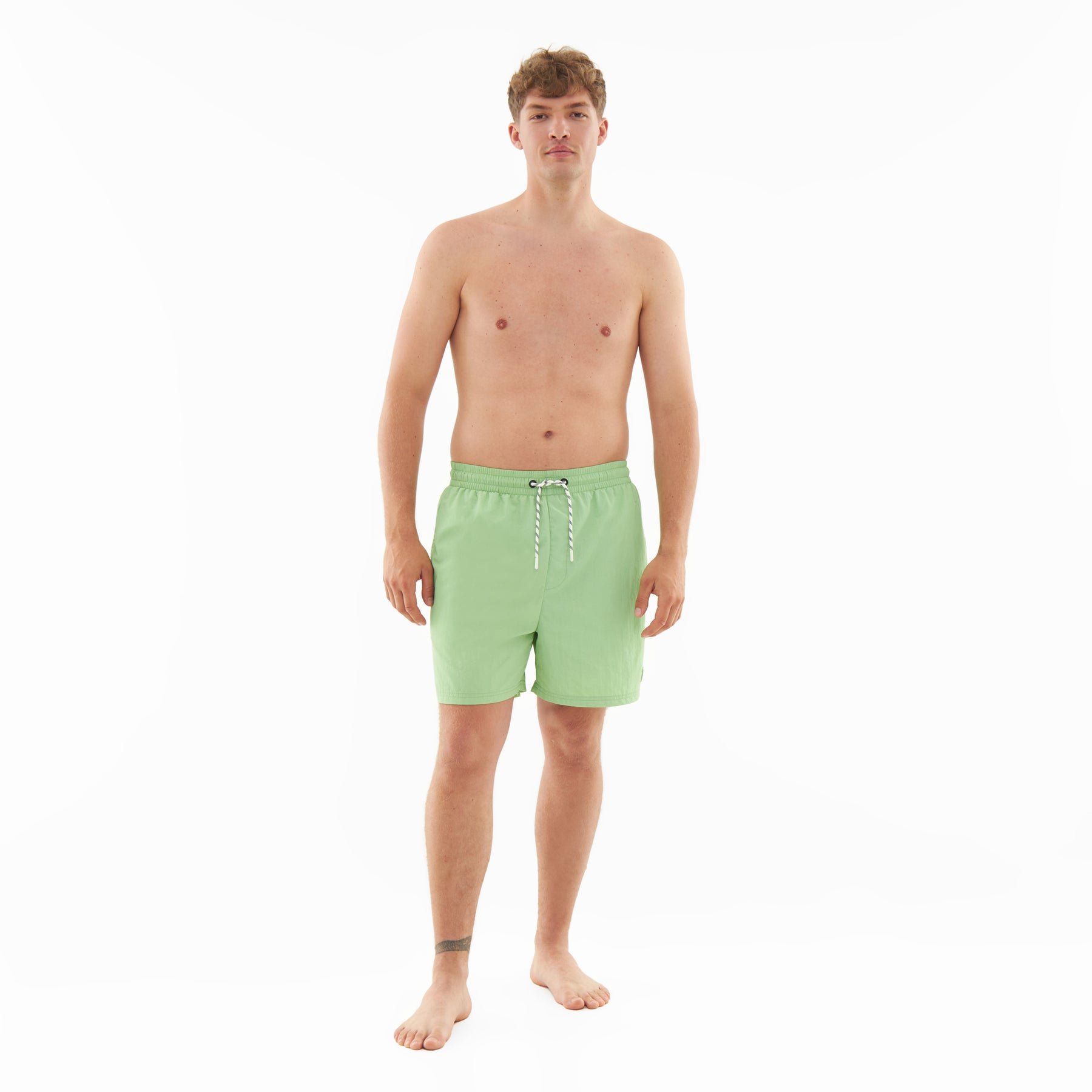 Mann trägt sage-grüne Badehose 'Jay' mit Kordelzug, steht barfuß vor weißem Hintergrund