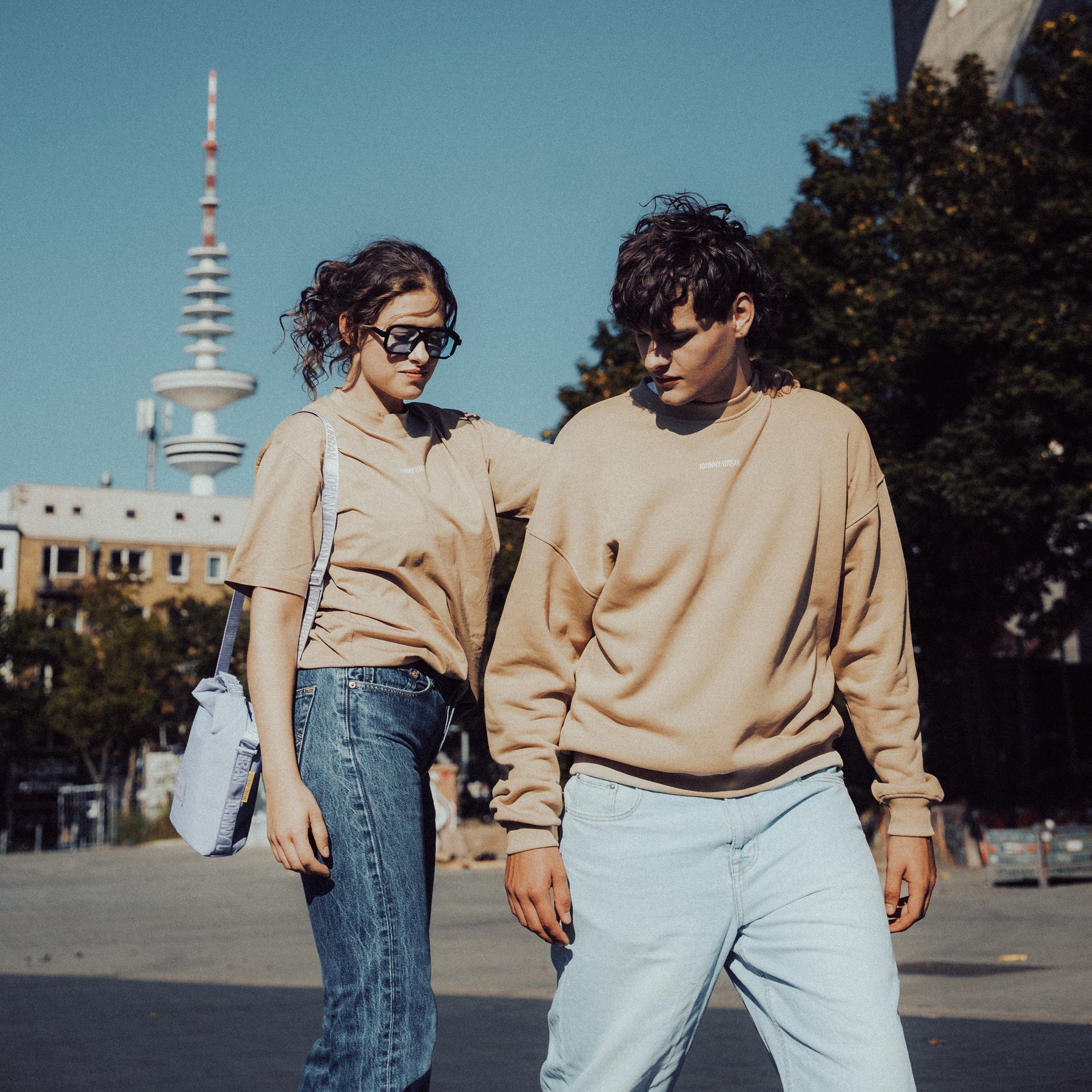 Frau in beige T-Shirt 'Sammy Johnny Urban' mit Jeans und Sonnenbrille, Mann in beige Pullover und hellen Jeans draußen in Stadtumgebung