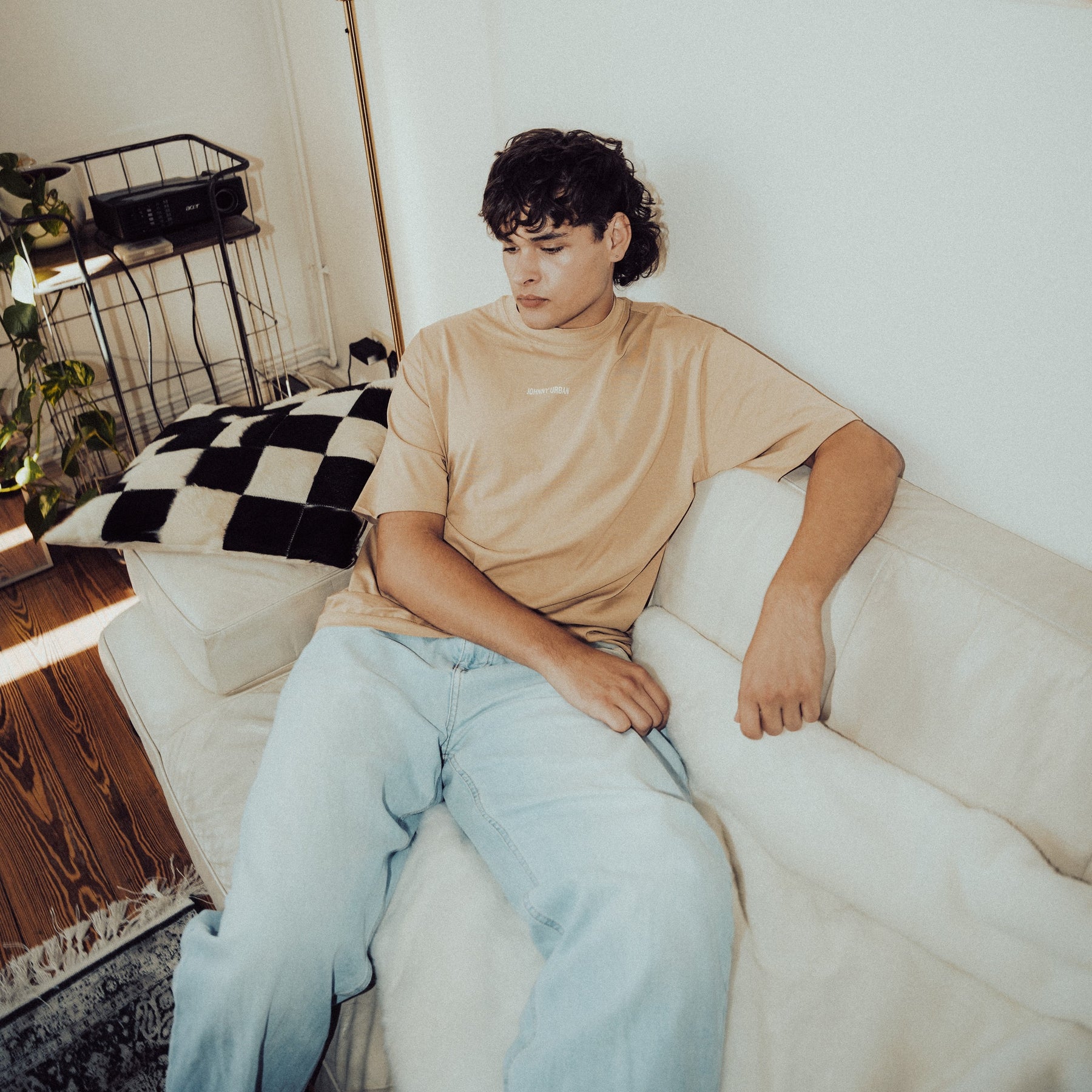 Mann sitzt auf weißem Sofa trägt oversized beige T-Shirt 'Sammy Johnny Urban' und helle Jeans