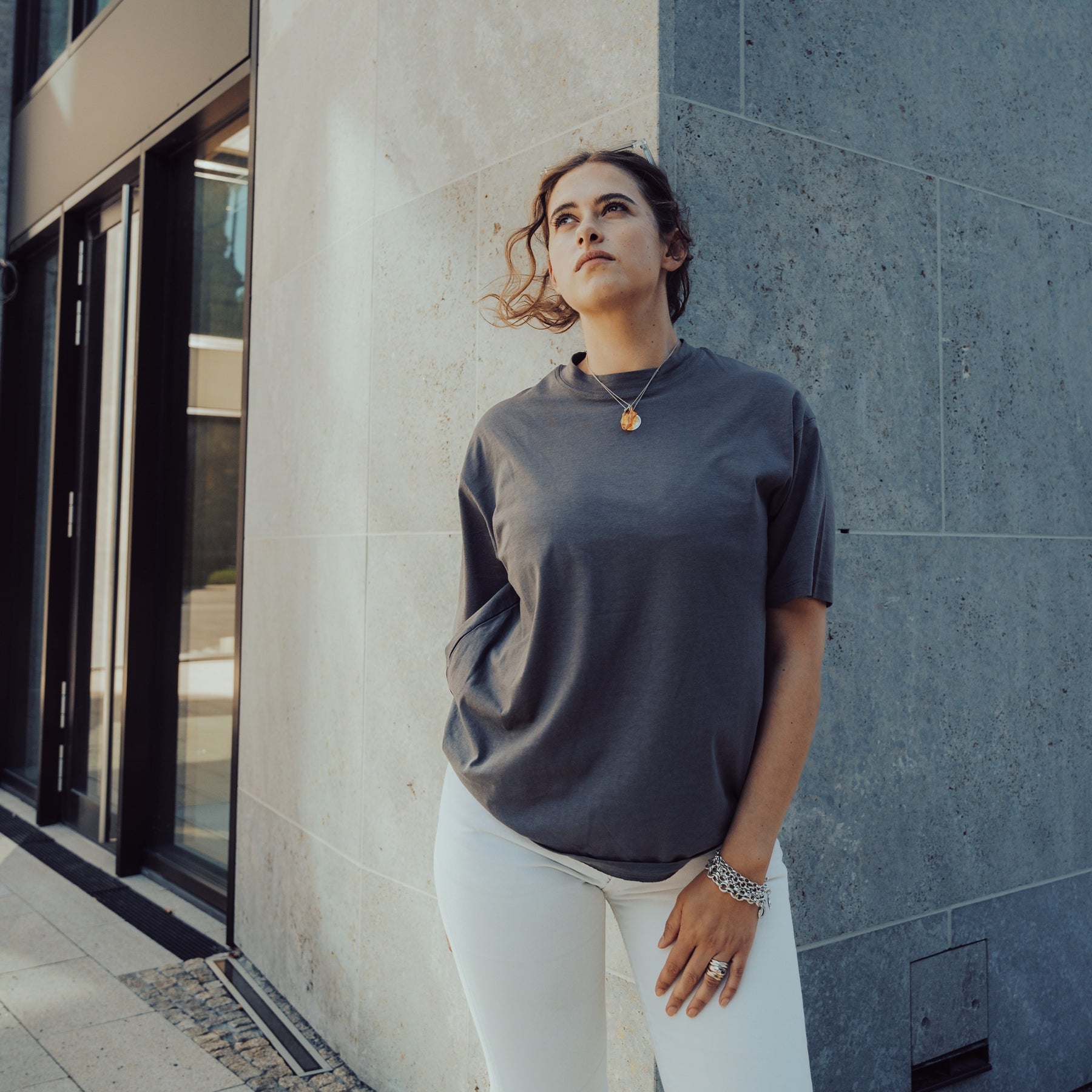 Frau trägt das graue Oversize T-Shirt 'Sammy' von Johnny Urban und weiße Hosen, steht vor einer grauen Steinwand