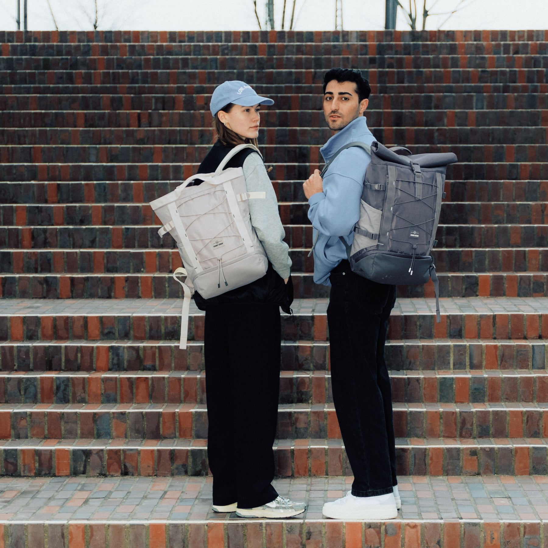 Zwei Personen auf einer Treppe tragen Rucksack 'Harper', eine beige Damenversion und eine schwarze Herrenversion