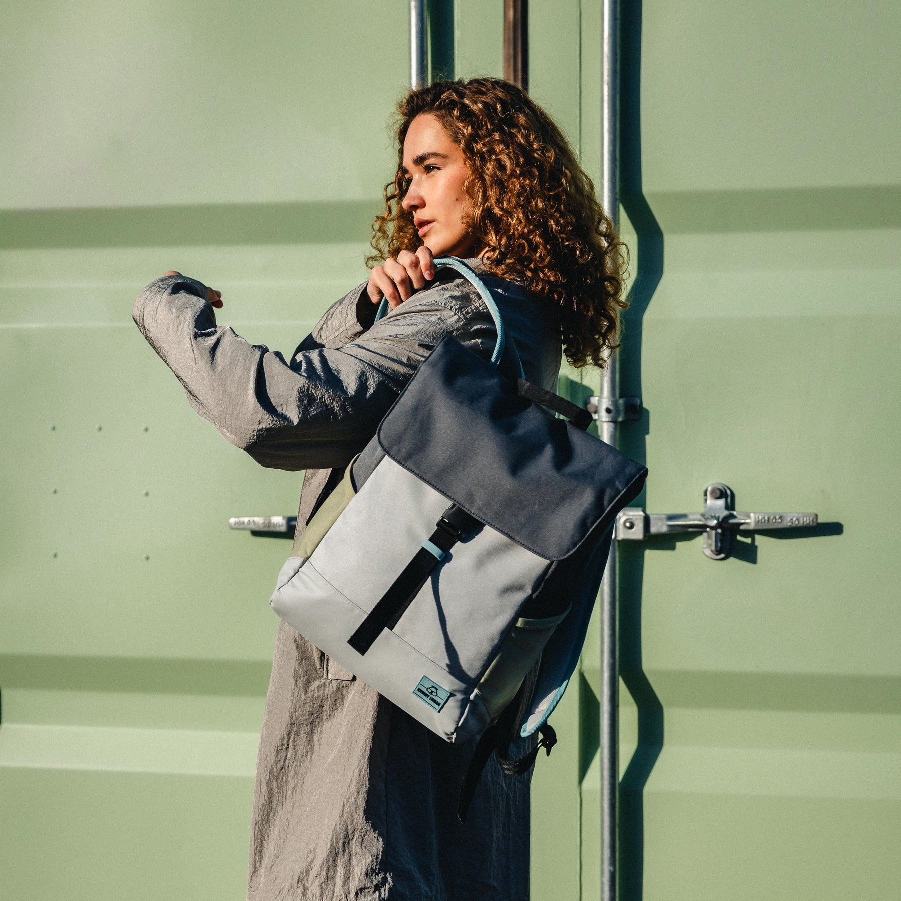 Frau mit lockigem Haar trägt den Tagesrucksack "Mika" in Grau und Grün über der Schulter vor grünem Hintergrund